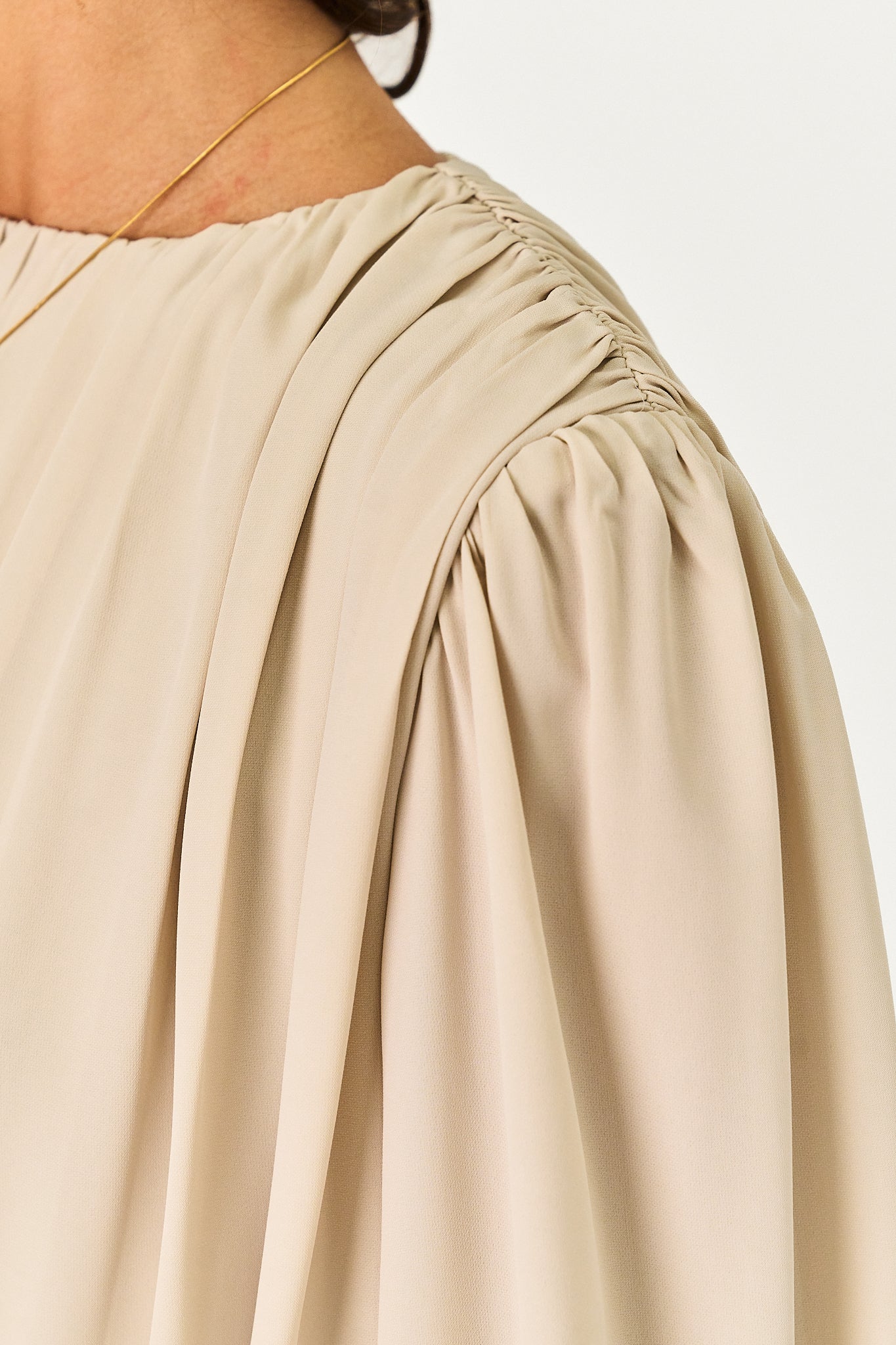 Close-up of a beige or light tan blouse with a gathered, smocked shoulder and long voluminous sleeves, worn by a model against a plain light background.