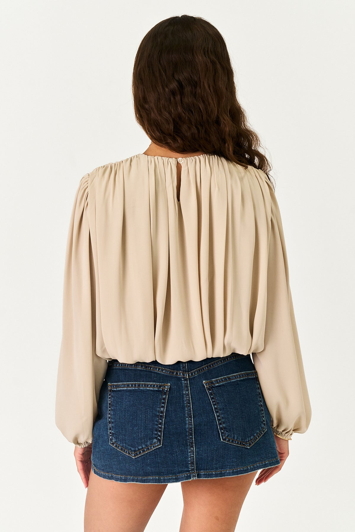 A woman shown from the back wearing a beige long-sleeve blouse with a gathered, pleated back and a blue denim mini skirt, against a plain white studio background.