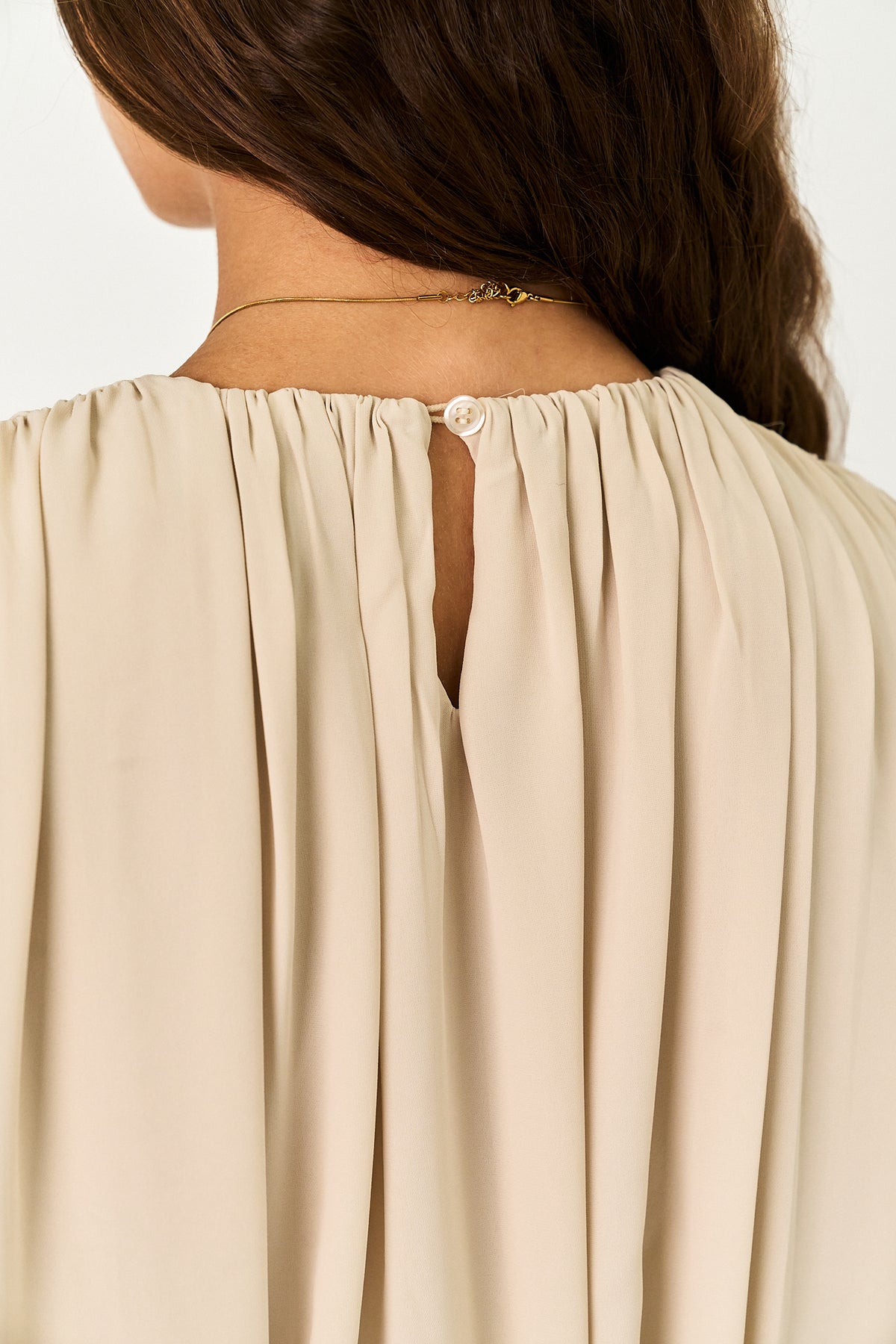 Close-up back view of a woman wearing a light beige pleated blouse with a small button closure at the center back and a thin gold necklace.