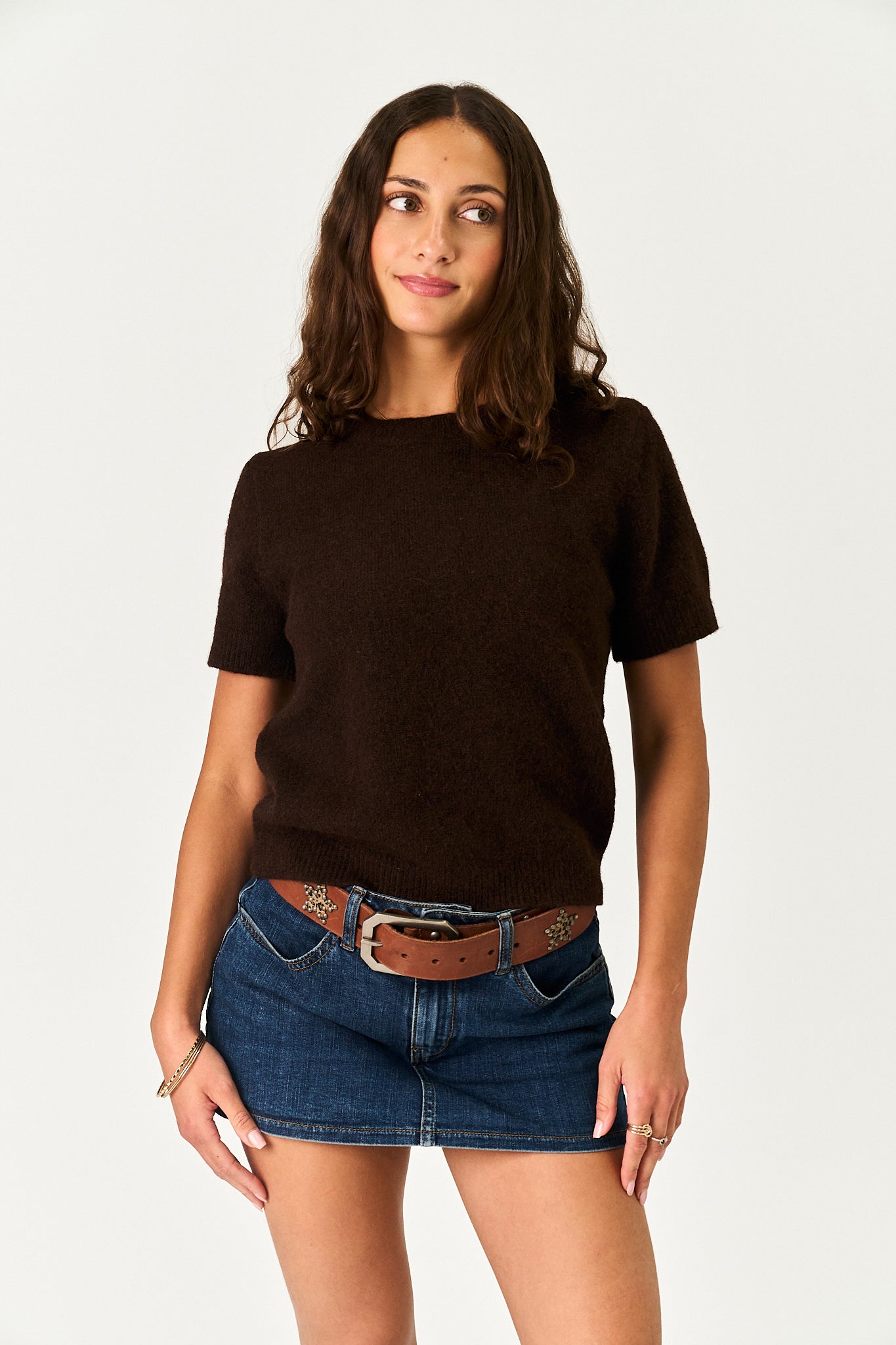 A woman modeling a dark brown short-sleeve knit top with blue denim shorts and a brown belt, photographed against a plain light gray studio background.