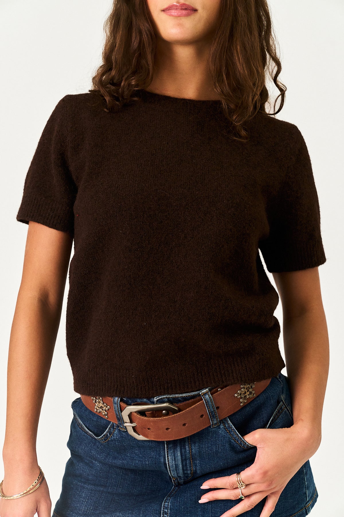 A woman wearing a dark brown short-sleeve knit sweater, styled with blue jeans and a brown leather belt, against a plain white background.