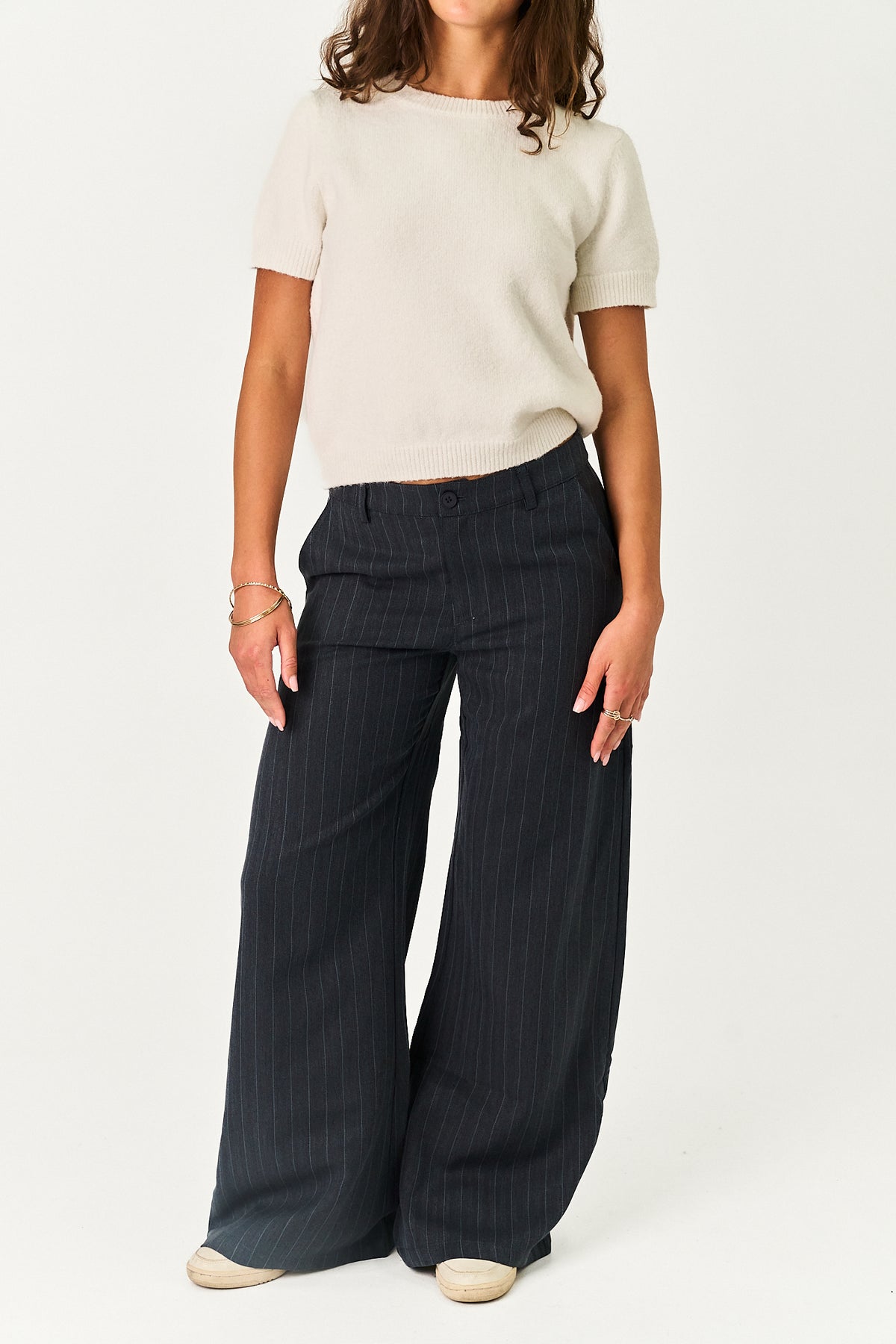 A model wearing a cream short-sleeve knit top and dark navy wide-leg pinstripe trousers, standing against a plain white studio background.