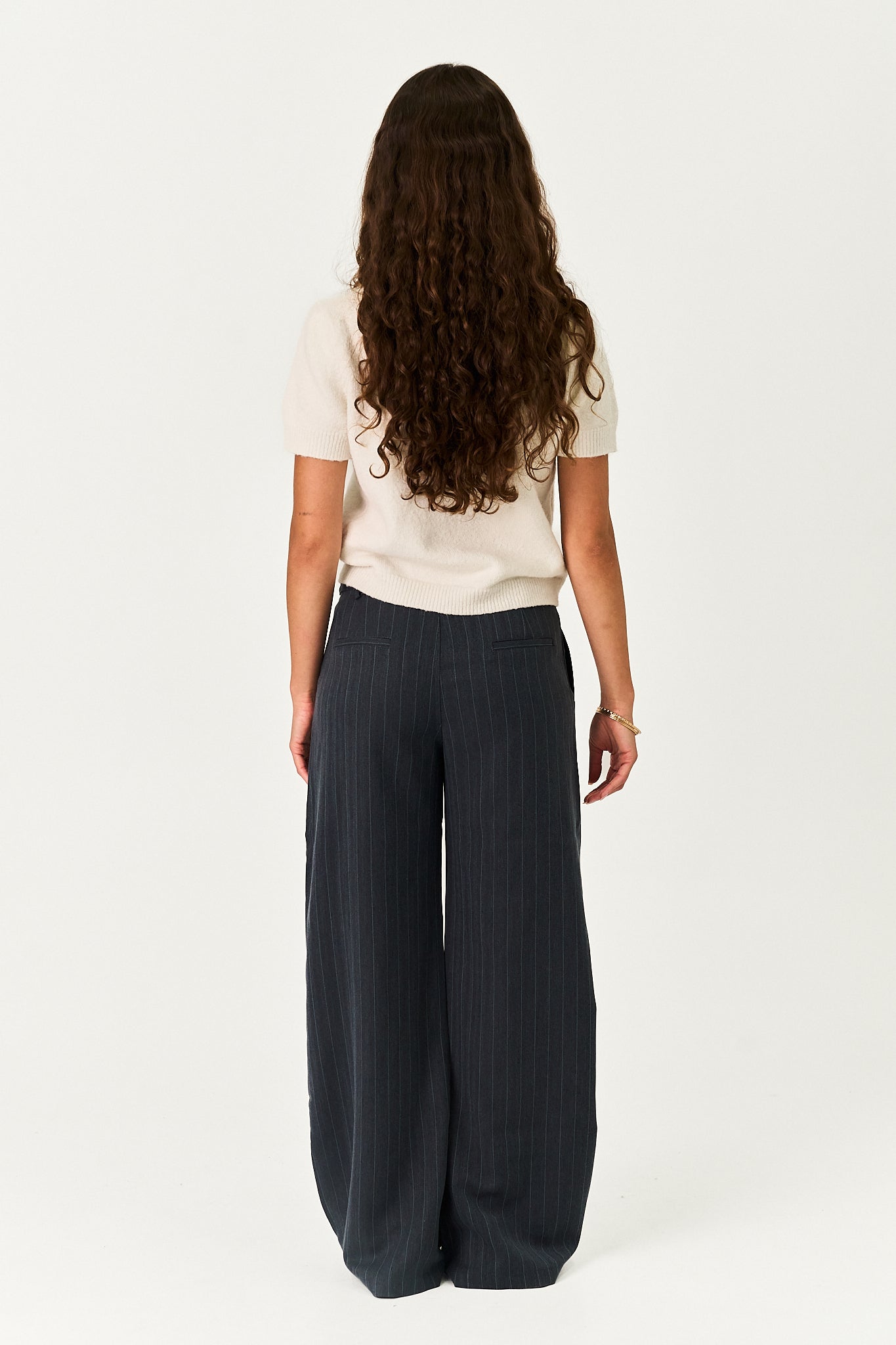A woman with long, curly brown hair is shown from behind wearing a cream short-sleeve t-shirt and dark navy wide-leg pinstripe trousers, standing against a plain white studio background.