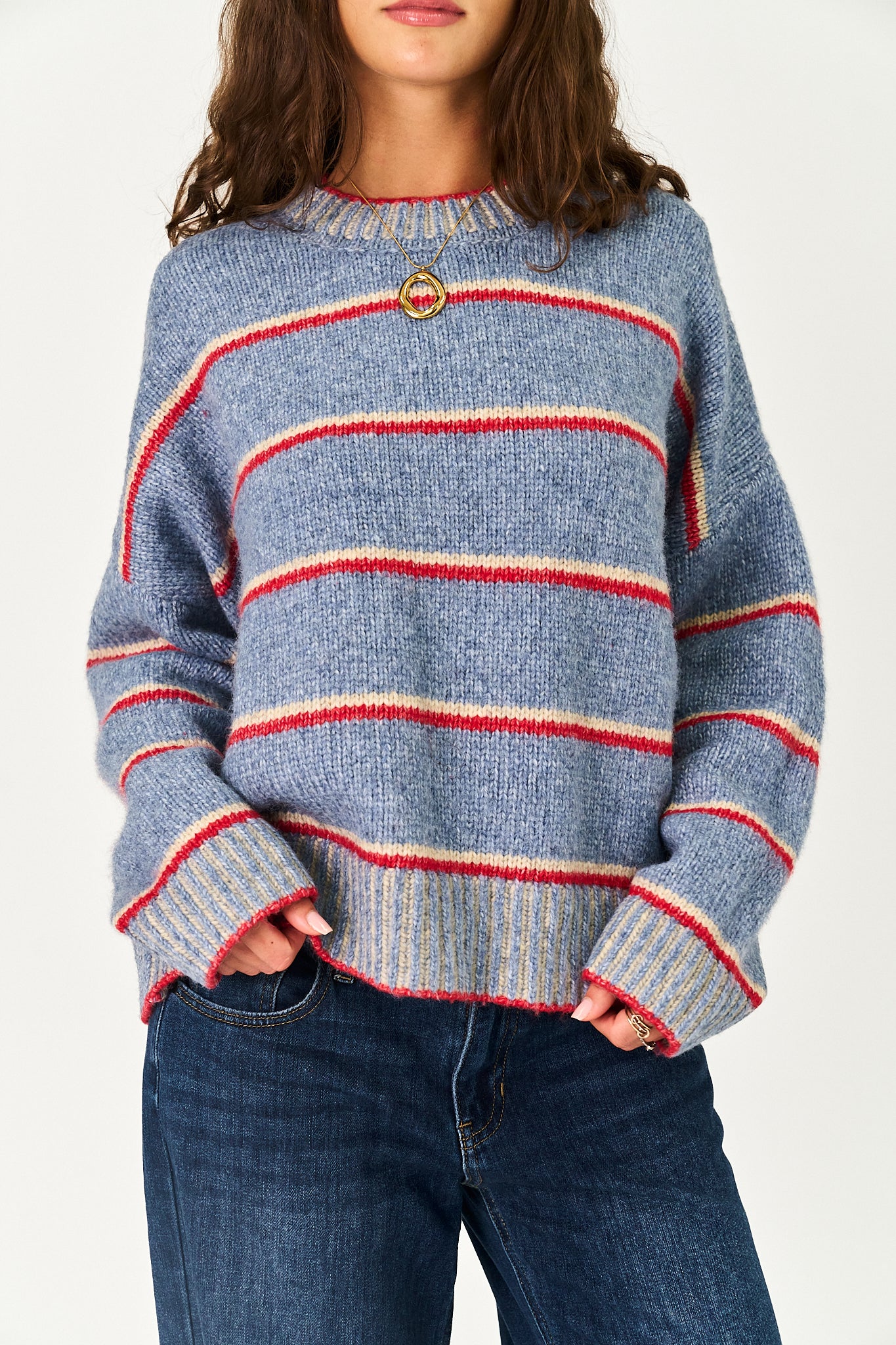 A woman wearing a blue knit sweater with horizontal red and cream stripes, paired with dark blue jeans, against a plain light background.