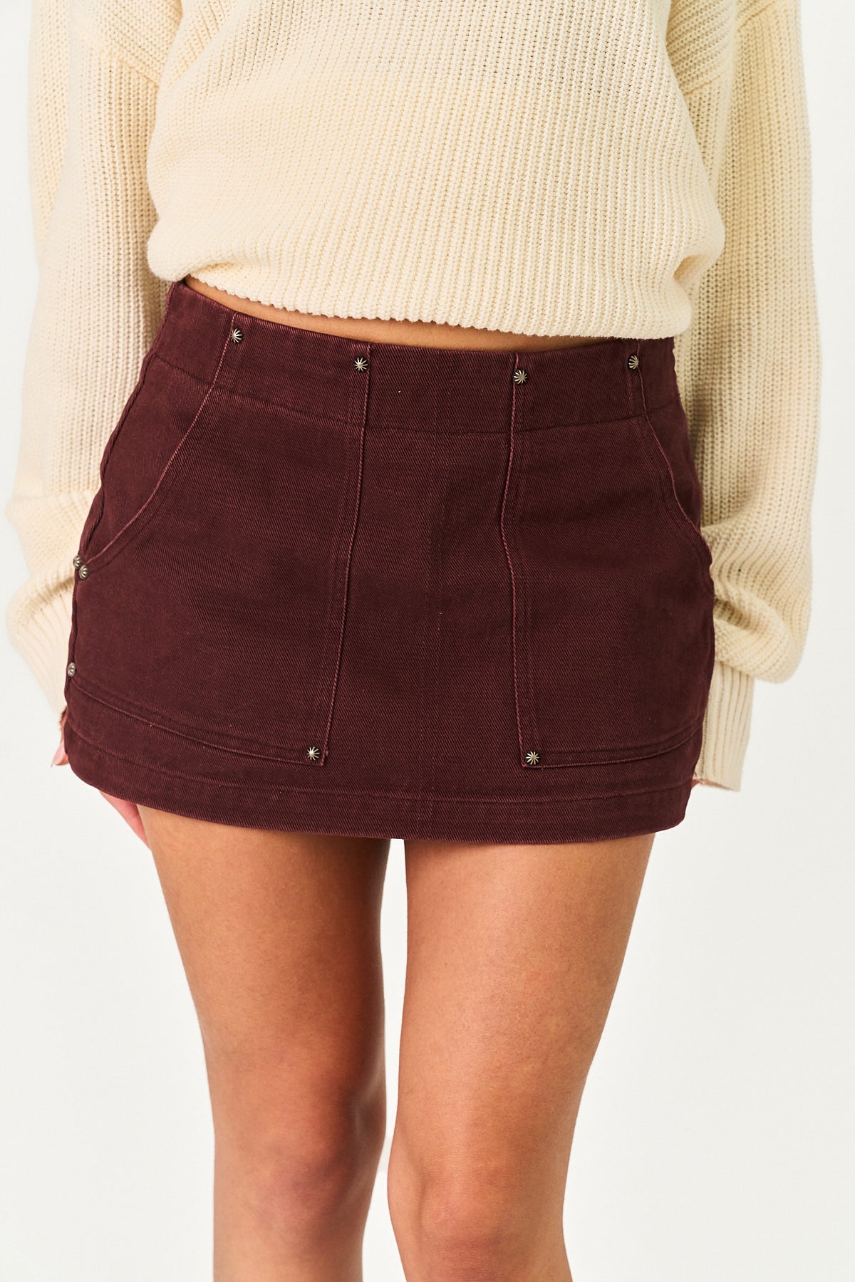 A close-up of a model wearing a dark burgundy mini skirt with silver rivet details and a cream ribbed knit sweater against a plain white background.