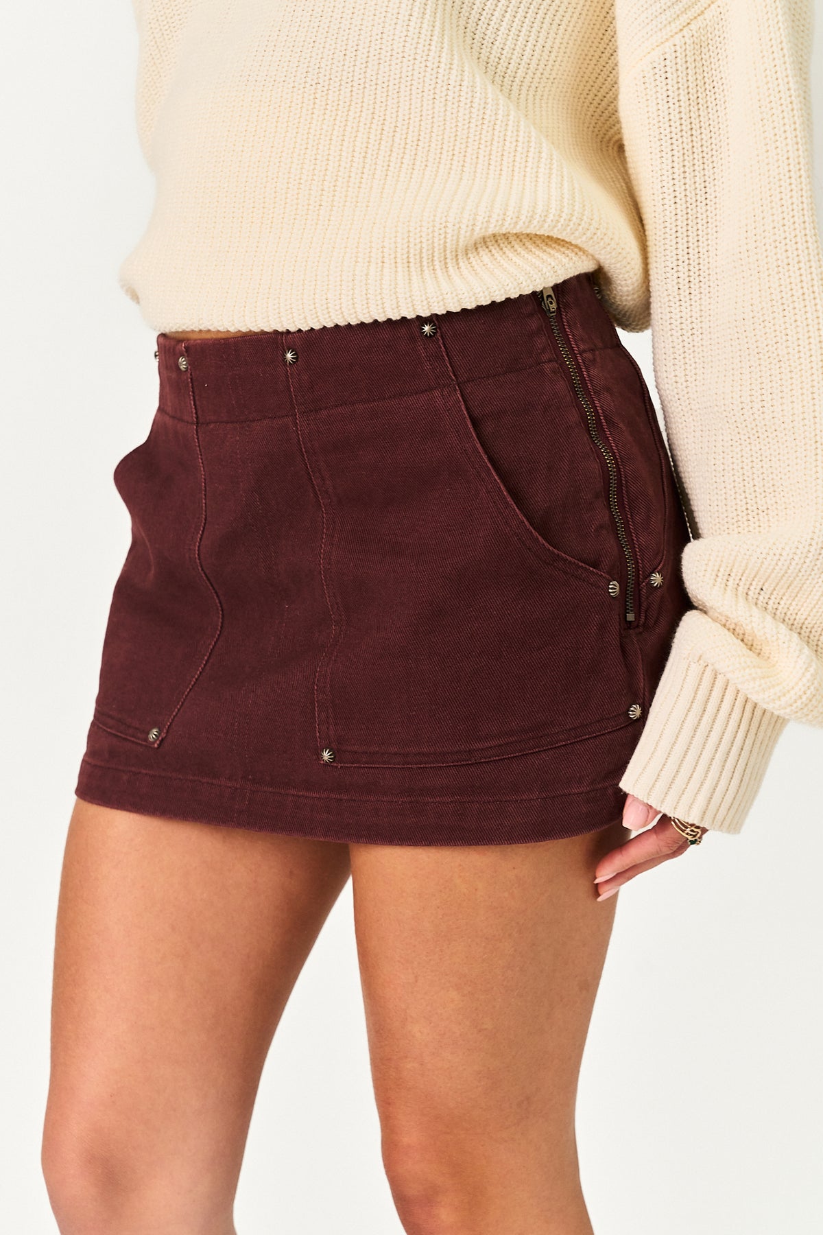 A close-up of a person wearing a dark burgundy mini skirt with front pockets, metal rivets, and side zipper details, paired with a cream ribbed sweater.