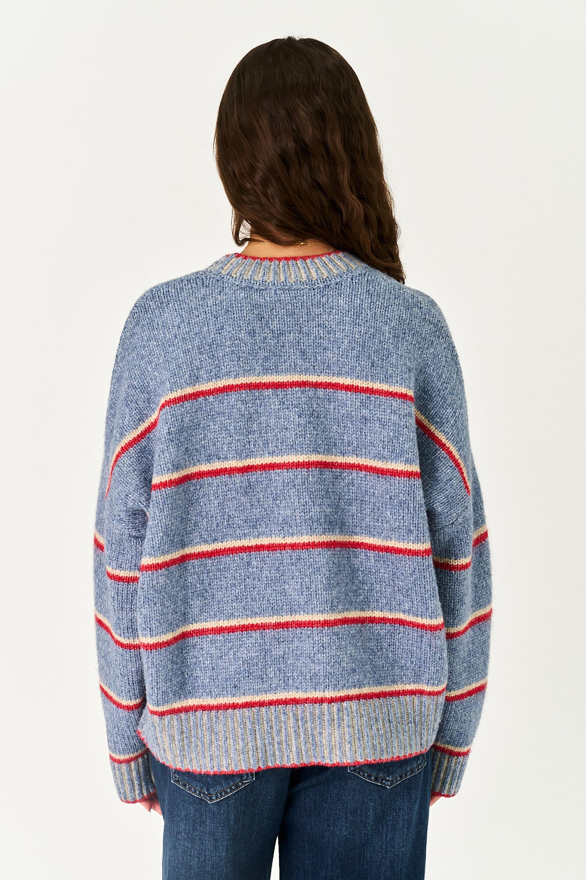 A woman viewed from behind wearing a blue knit sweater with horizontal red and cream stripes, paired with blue jeans, against a plain off-white background.