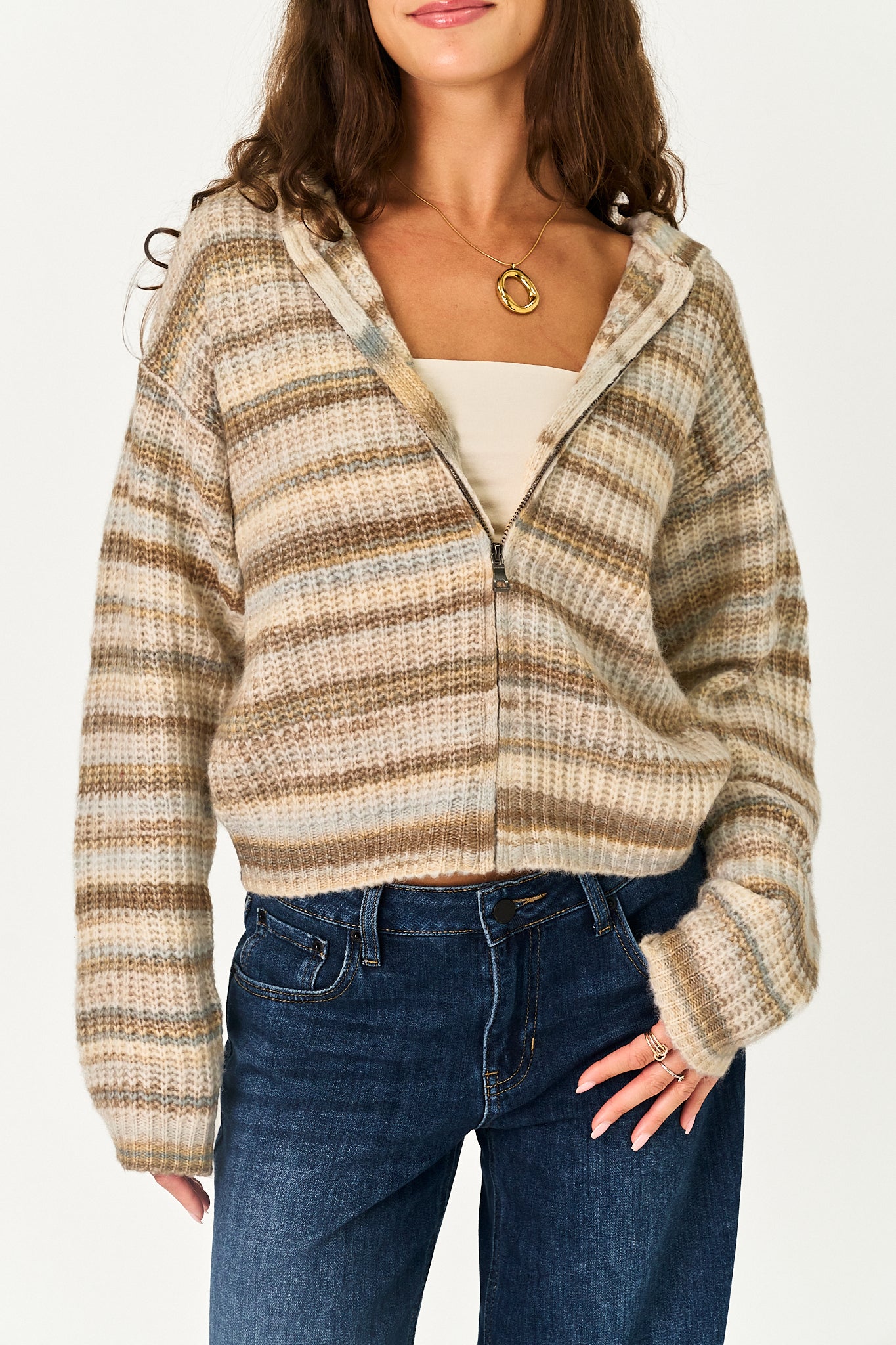 A woman wearing a cropped zip-up hooded sweater with horizontal beige, brown, and tan stripes, styled over a cream top and blue jeans.