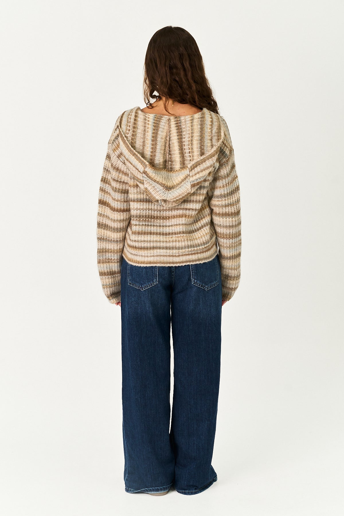 A woman viewed from behind wearing a beige and brown horizontally striped knit hooded sweater and dark blue wide-leg jeans, standing against a plain white studio background.