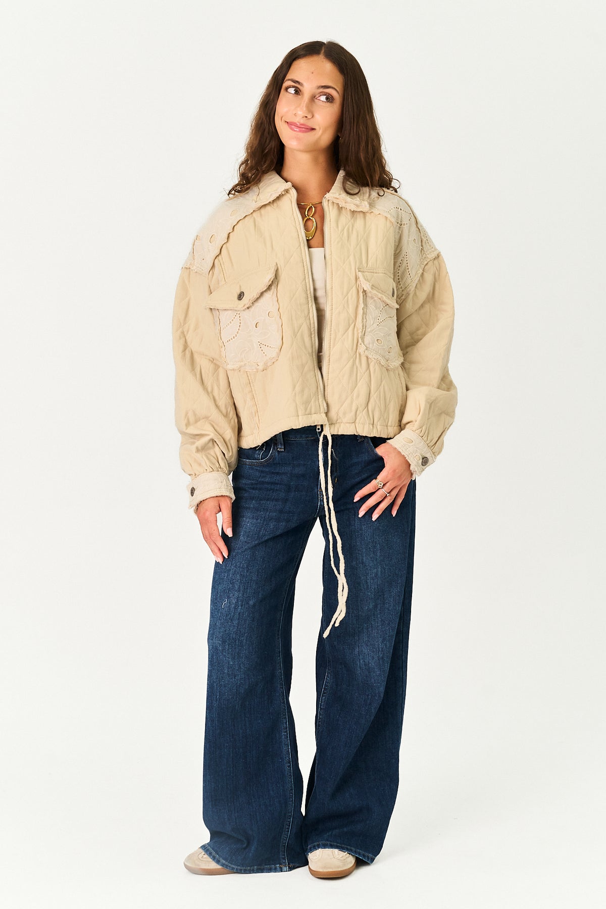 A woman modeling a cropped beige quilted jacket with front flap pockets, paired with dark blue wide-leg jeans and beige shoes, against a plain white studio background.