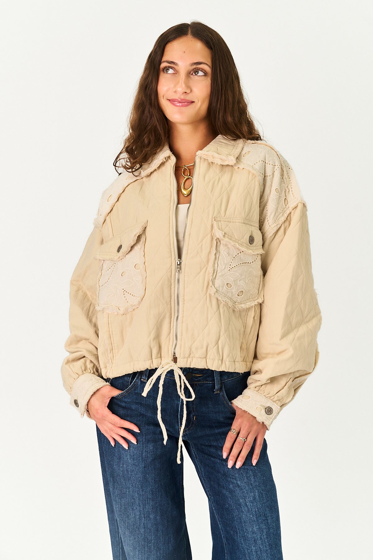 A woman modeling a beige quilted jacket with lace applique details, paired with blue jeans, against a plain white studio background.