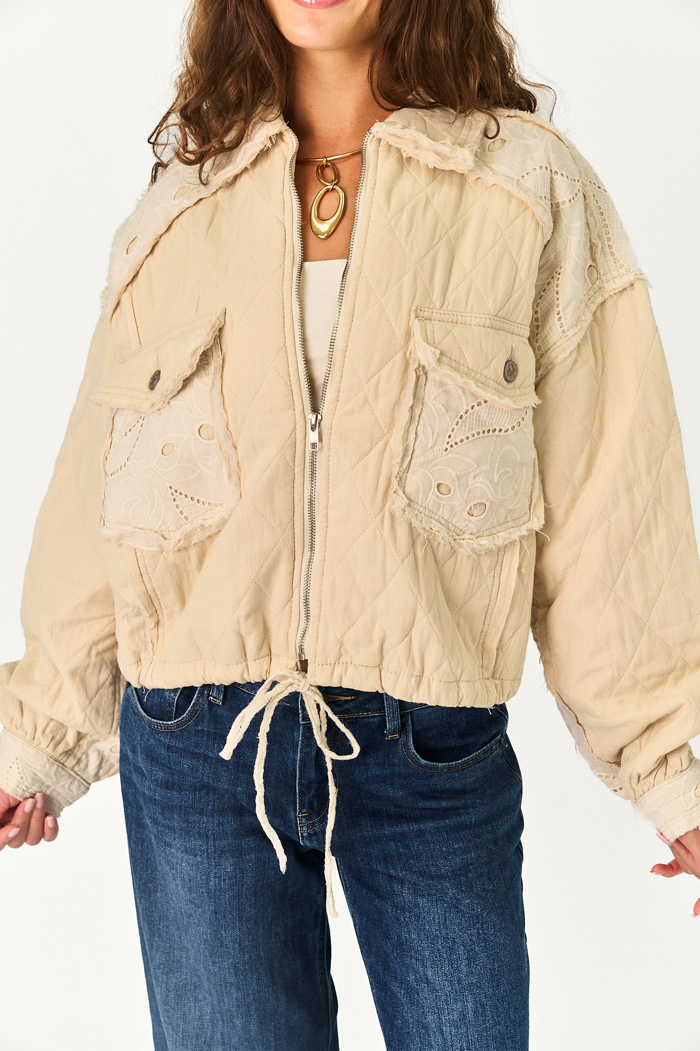 A woman wearing a cropped beige quilted jacket with lace applique details, front pockets, and a drawstring hem, paired with blue jeans against a plain white background.