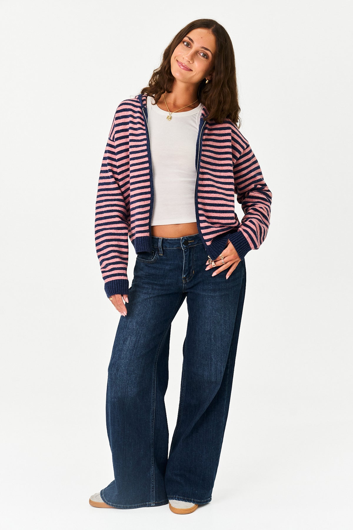 A woman modeling a navy and pink striped zip-up hoodie over a white crop top and dark blue wide-leg jeans, standing against a plain white studio background.