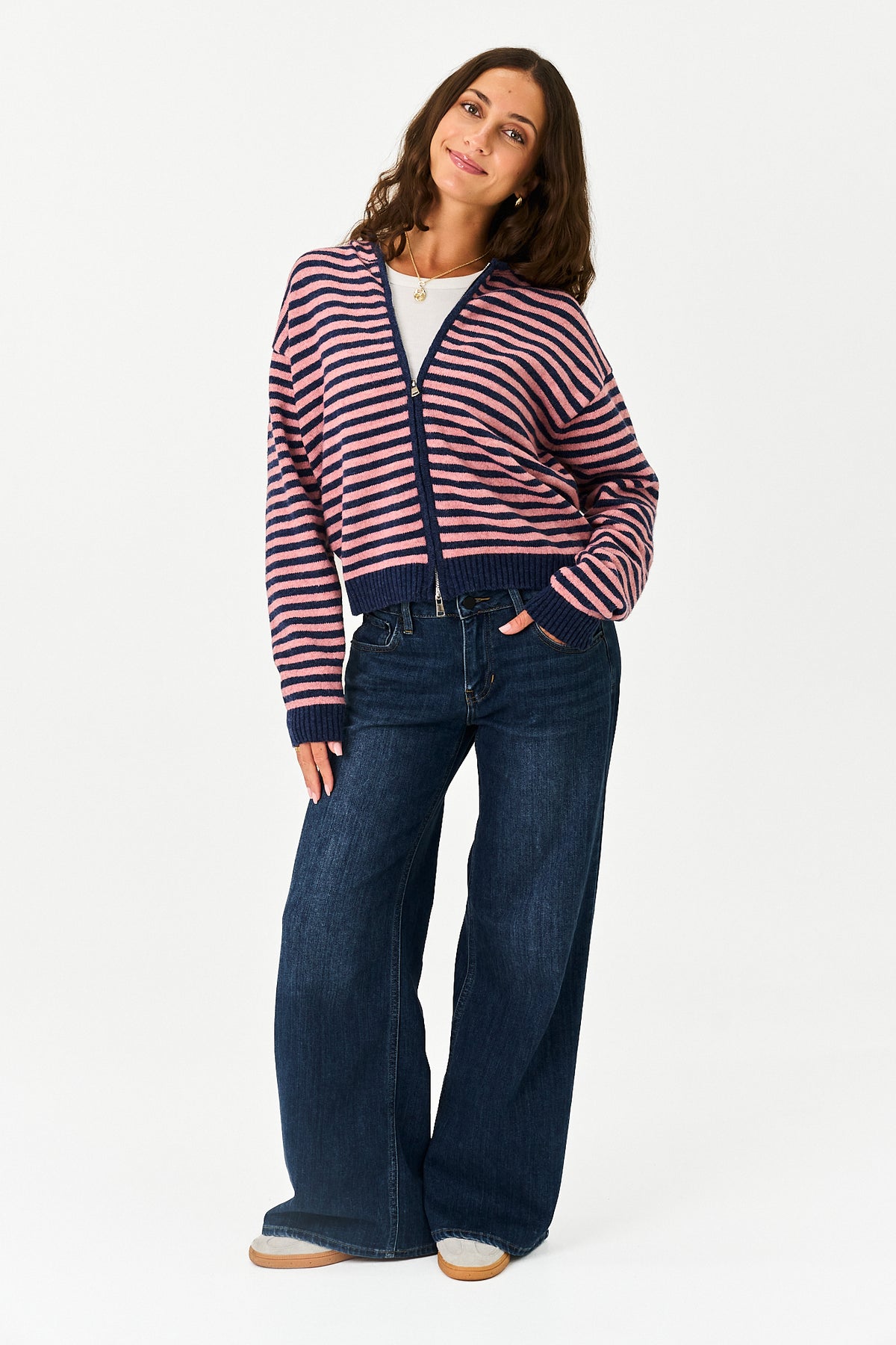 A woman modeling a pink and navy blue horizontally striped zip-up cardigan sweater, paired with dark blue wide-leg jeans, against a plain white studio background.