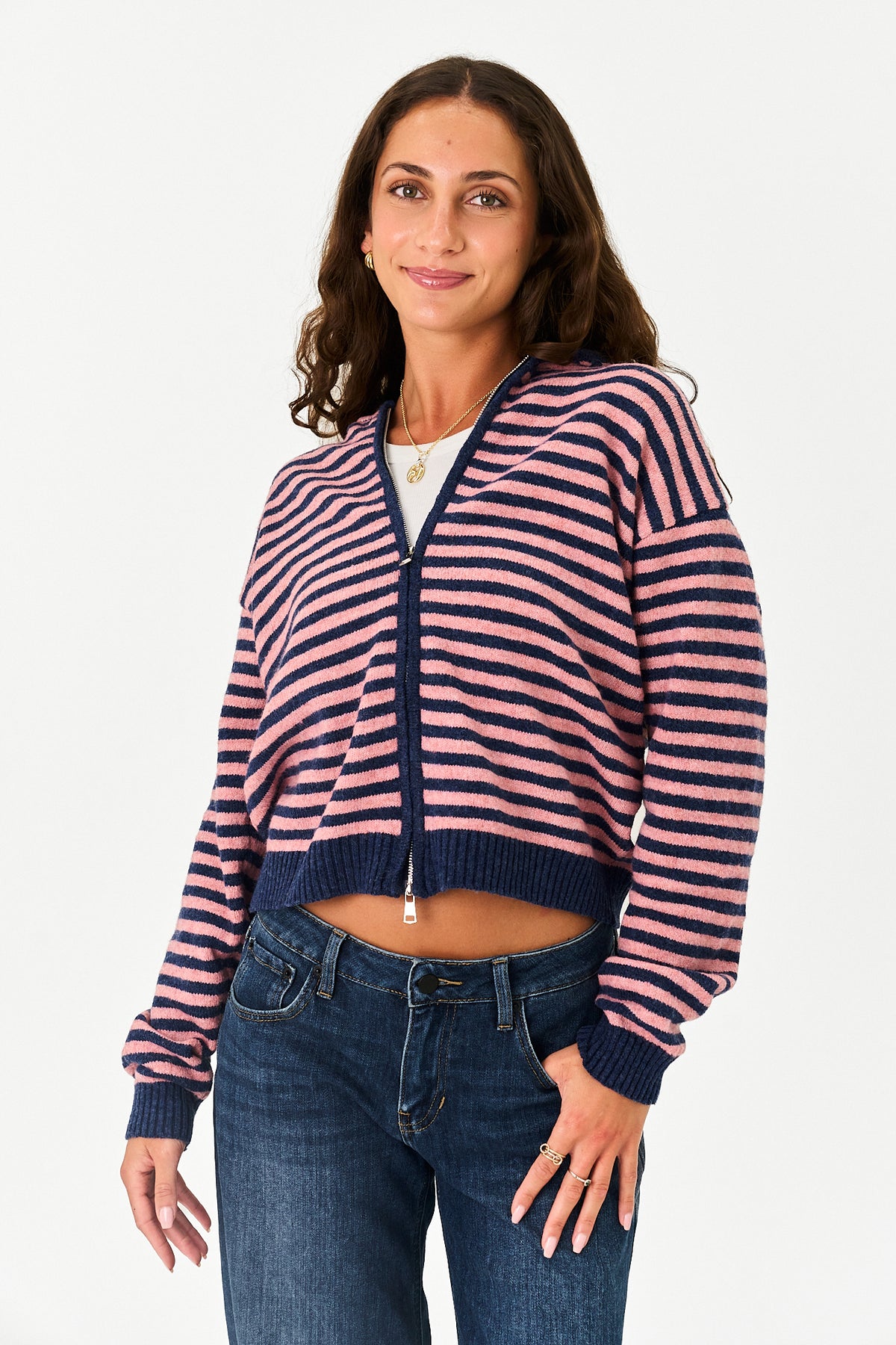 A woman modeling a cropped, long-sleeve zip-up knit cardigan with horizontal navy blue and pink stripes, paired with dark blue jeans against a plain white studio background.