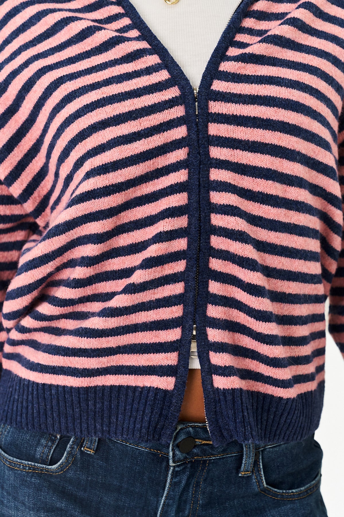 A close-up of a person wearing a pink and navy blue horizontally striped knit cardigan with a front zipper, paired with blue jeans.