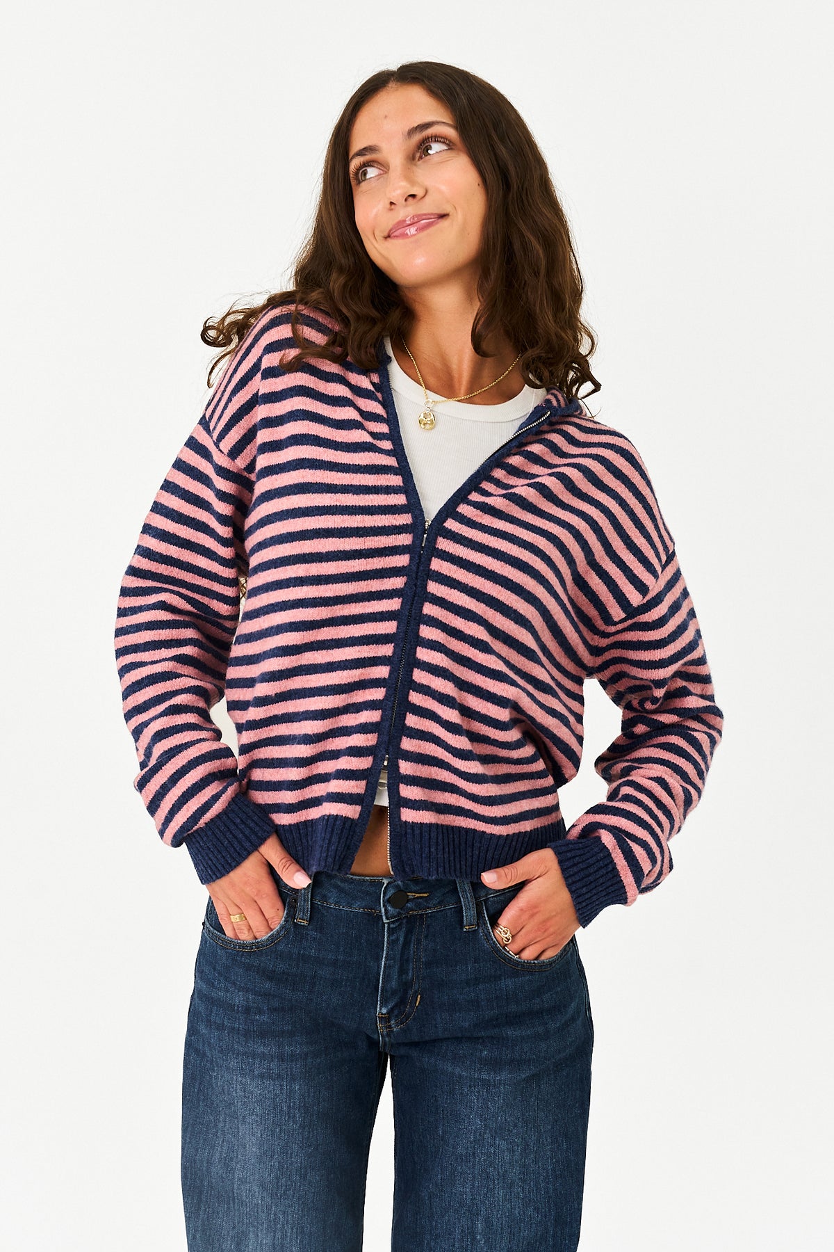 A woman wearing a pink and navy blue horizontally striped zip-up hoodie with pockets, paired with a white t-shirt and blue jeans, against a plain white background.