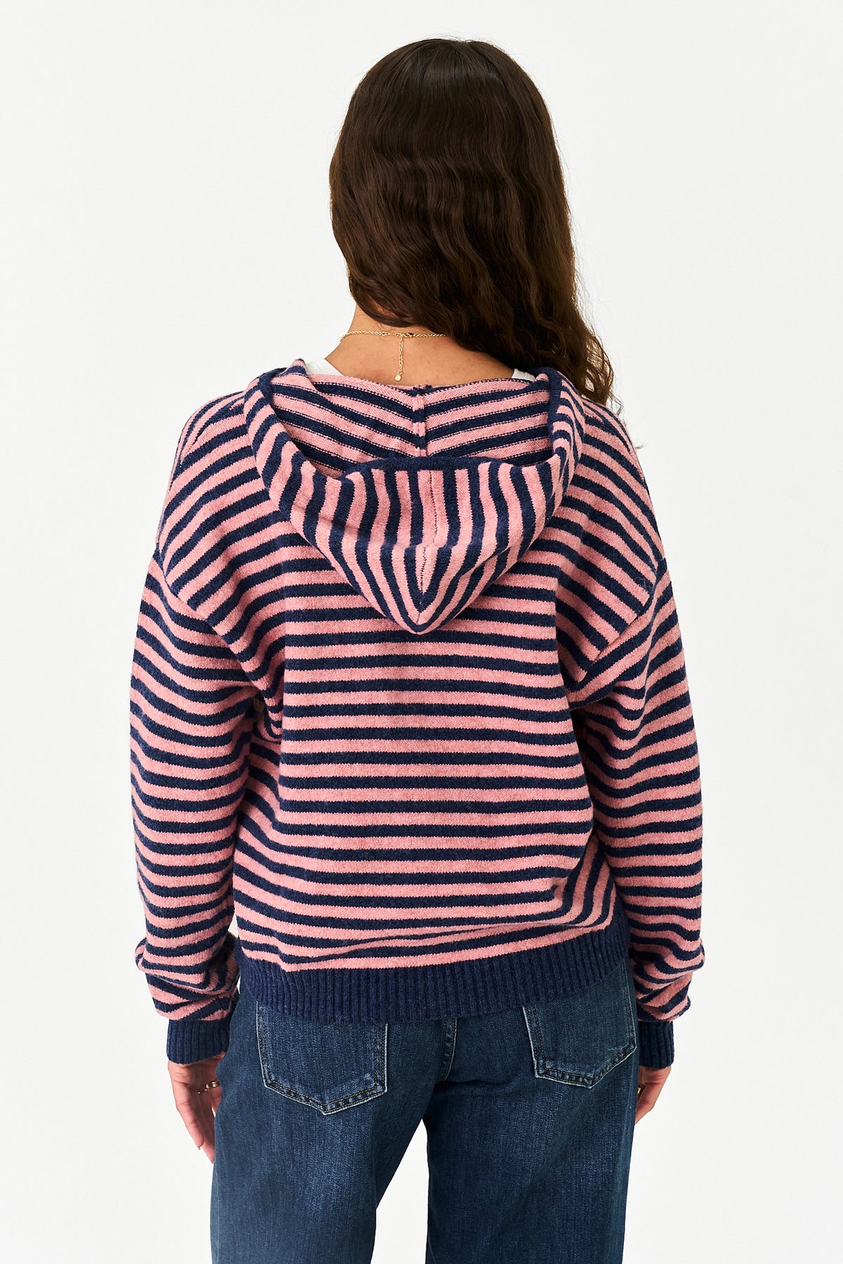 A woman viewed from behind wearing a pink and navy blue horizontally striped knit hoodie with a ribbed hem and cuffs, paired with blue jeans, against a plain white background.