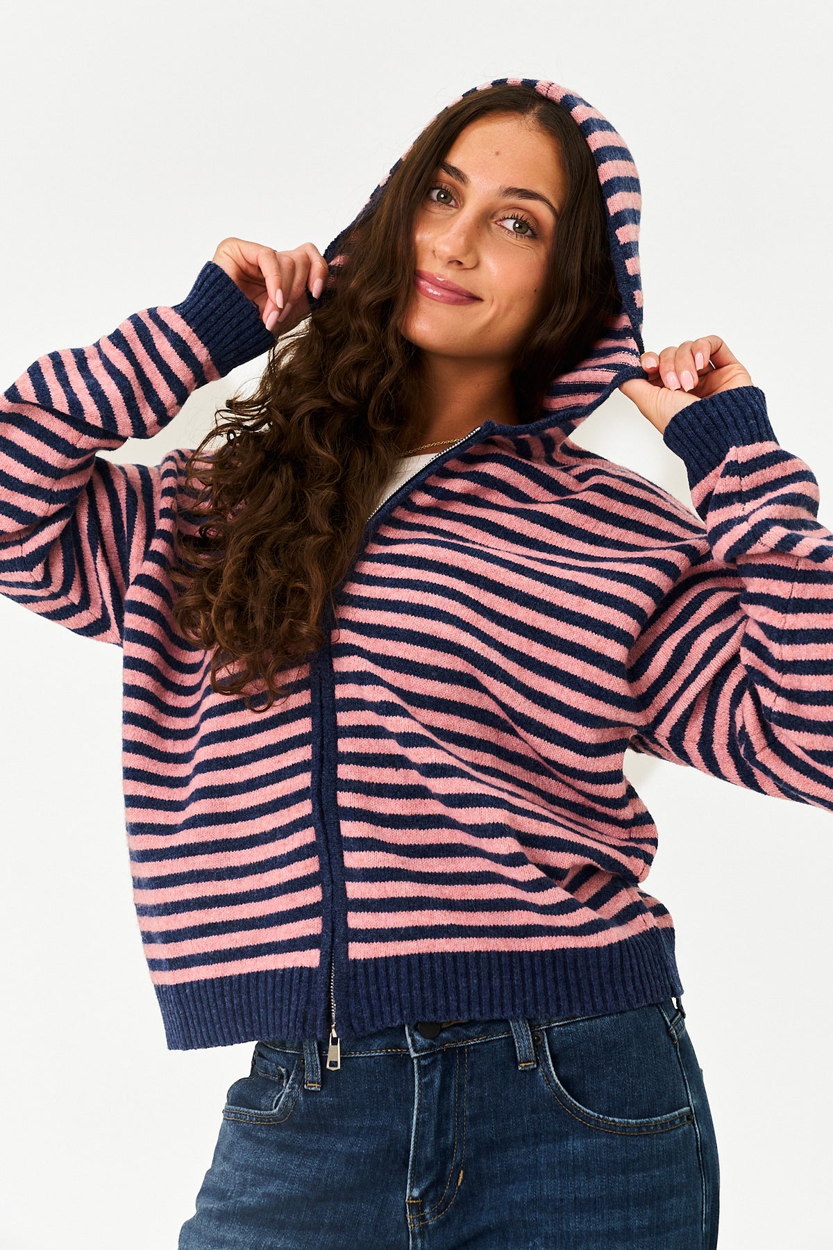 A woman wearing a pink and navy blue striped zip-up hoodie with a ribbed hem, cuffs, and hood, paired with blue jeans, against a plain white background.