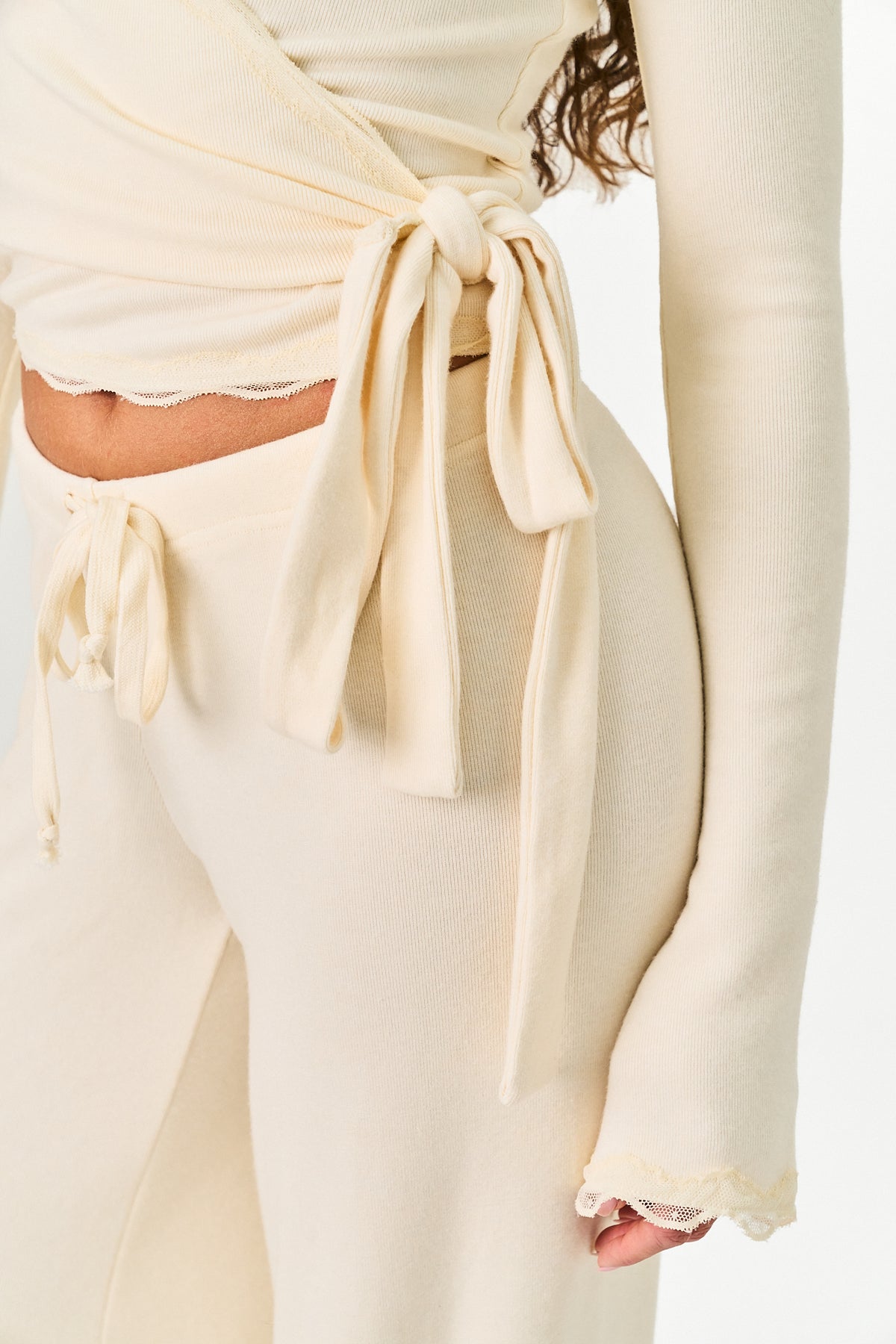 Cream-colored knit loungewear set featuring a long-sleeve crop top with a side tie and matching high-waisted drawstring pants, shown on a model against a white background.