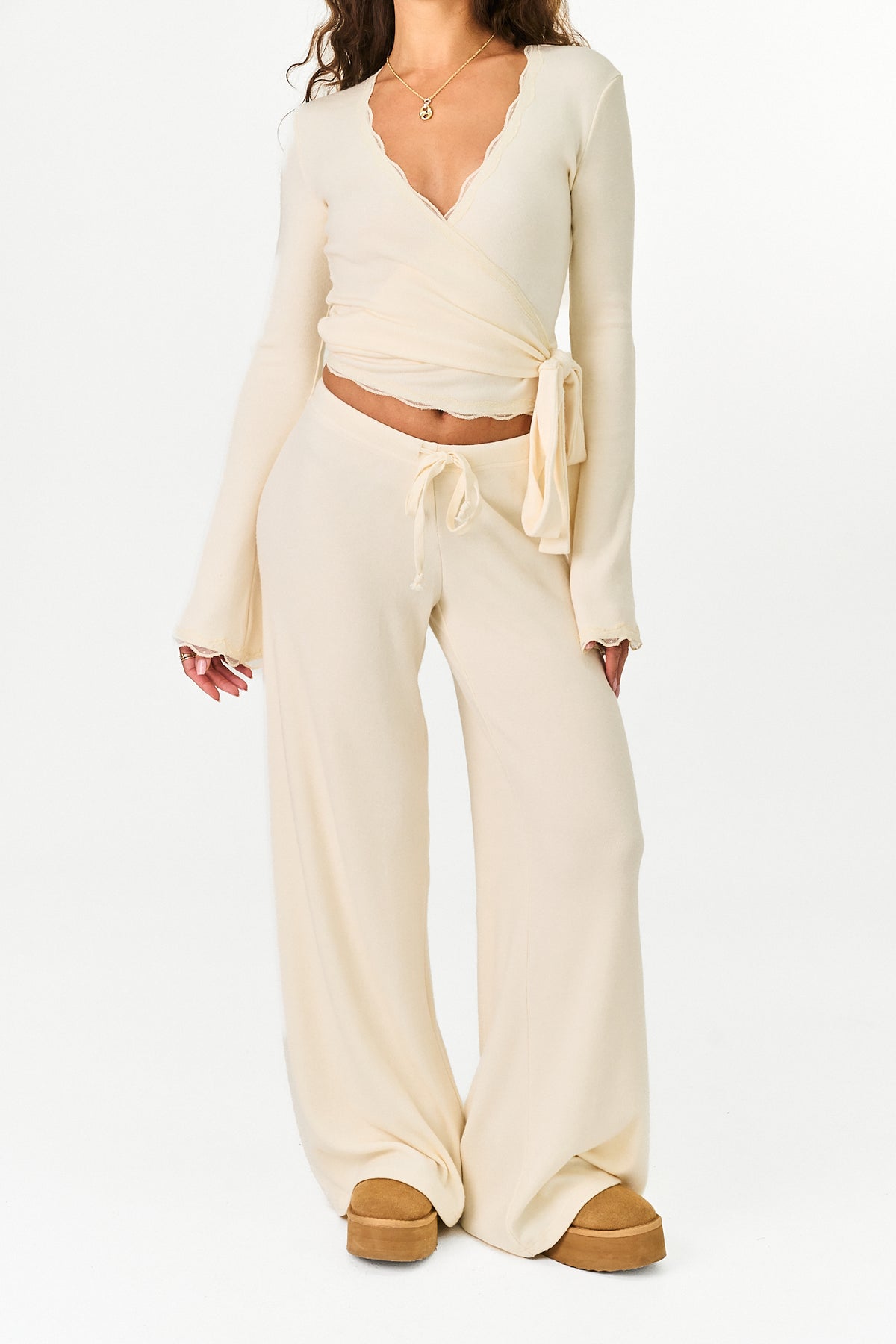 A model wearing a cream-colored two-piece knit set consisting of a long-sleeve wrap top and matching wide-leg pants, paired with brown platform shoes against a white studio background.
