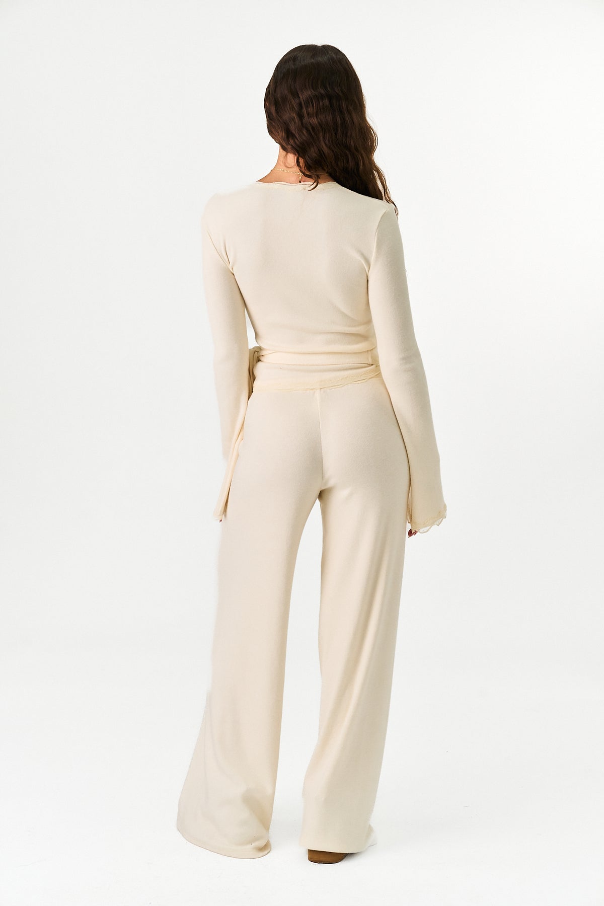 A woman viewed from the back wearing a cream-colored long-sleeve jumpsuit with a wrap-style waist and wide-leg pants, set against a plain white studio background.