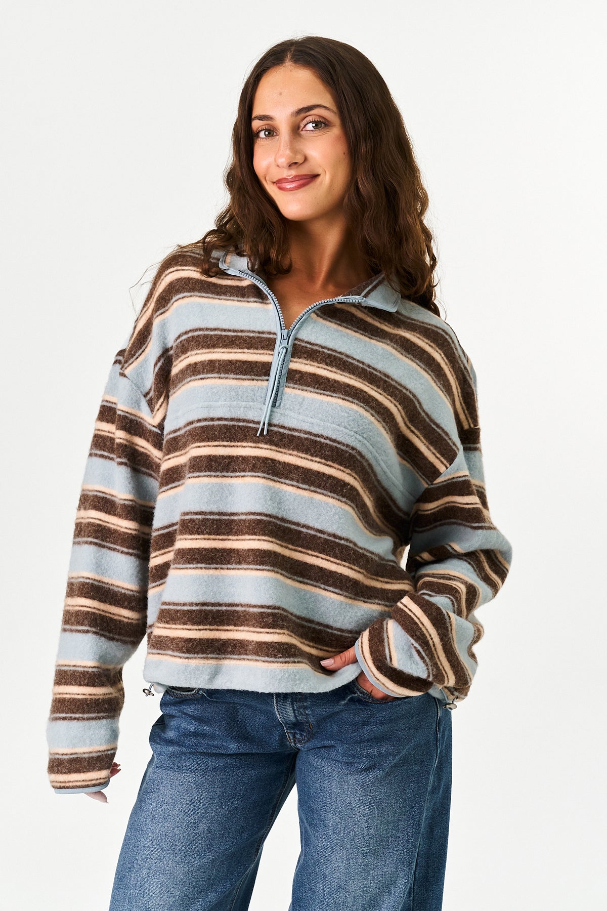 A woman wearing a fuzzy, long-sleeve quarter-zip pullover sweater with horizontal stripes in brown, beige, and light blue, paired with blue jeans, against a plain white studio background.