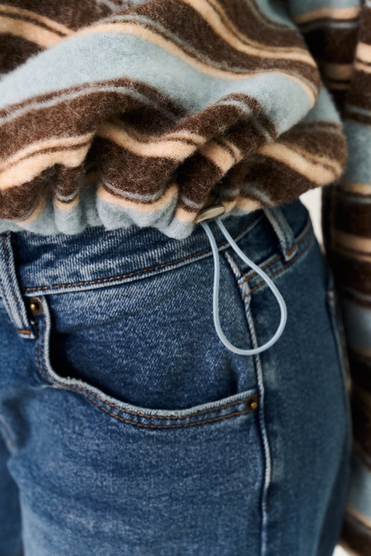 Close-up of a person wearing a brown, beige, and light blue striped knit top tucked into blue jeans, with a light blue drawstring visible at the waist.