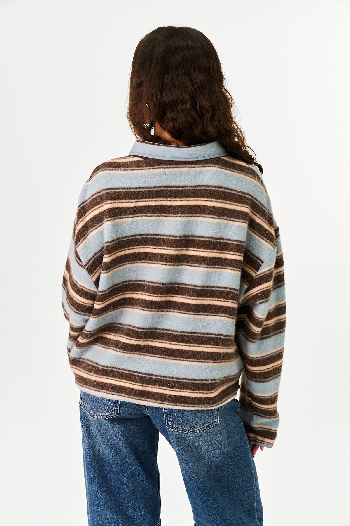A person shown from the back wearing a striped knit sweater with horizontal bands of light blue, brown, and beige, paired with blue jeans, against a plain white studio background.