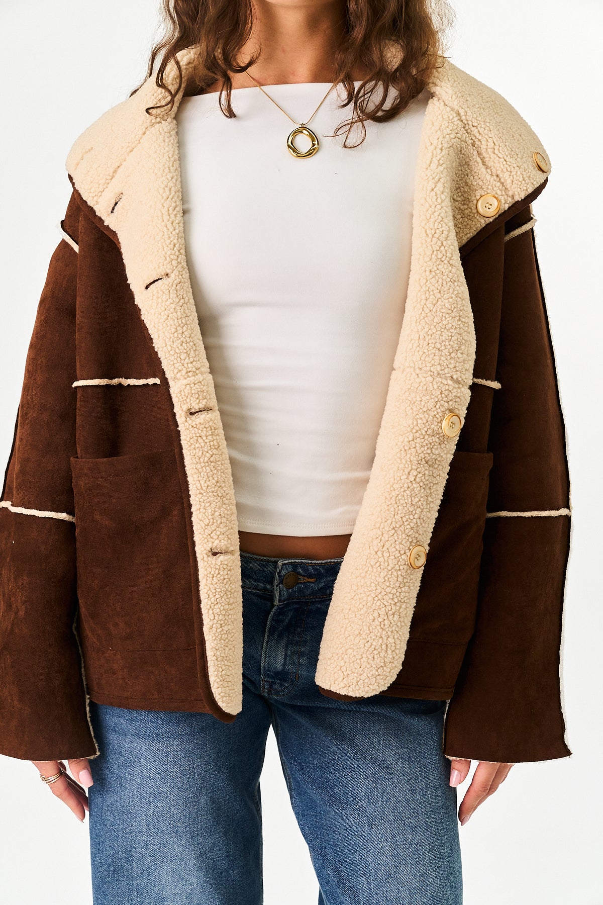A person wearing a brown suede-like jacket with a cream sherpa lining over a white top and blue jeans, shown from the shoulders to the upper thighs against a plain light background.