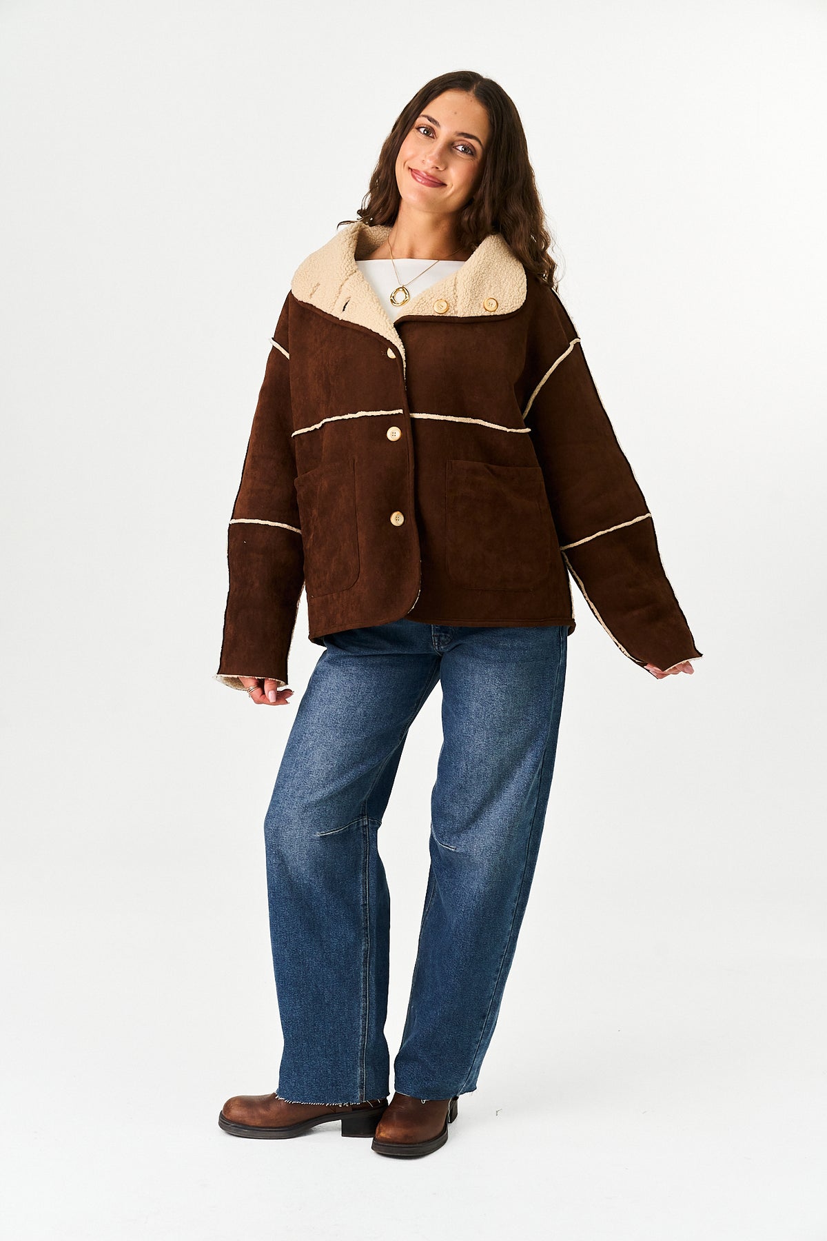 A person wearing a brown suede-like jacket with a light beige sherpa collar and contrast beige piping, paired with blue jeans, against a plain white studio background.