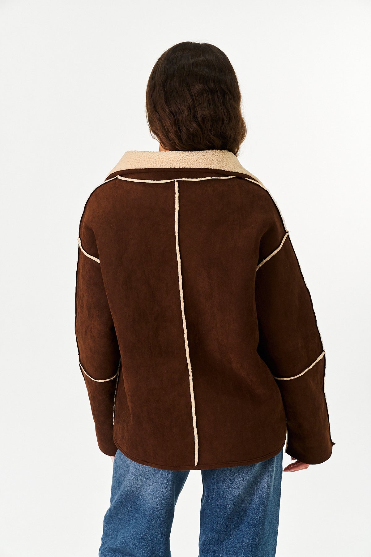 A person shown from the back wearing a brown suede-like jacket with a light beige sherpa collar and contrast beige piping, paired with blue jeans, against a plain white studio background.