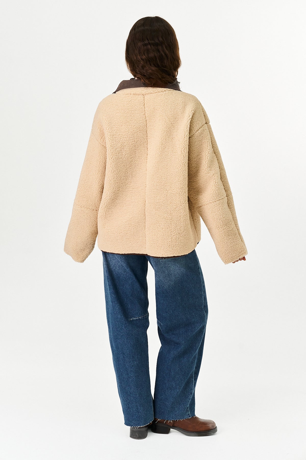 A person wearing a light brown sherpa-like jacket with a brown suede collar and contrast beige piping, paired with blue jeans, against a plain white studio background.
