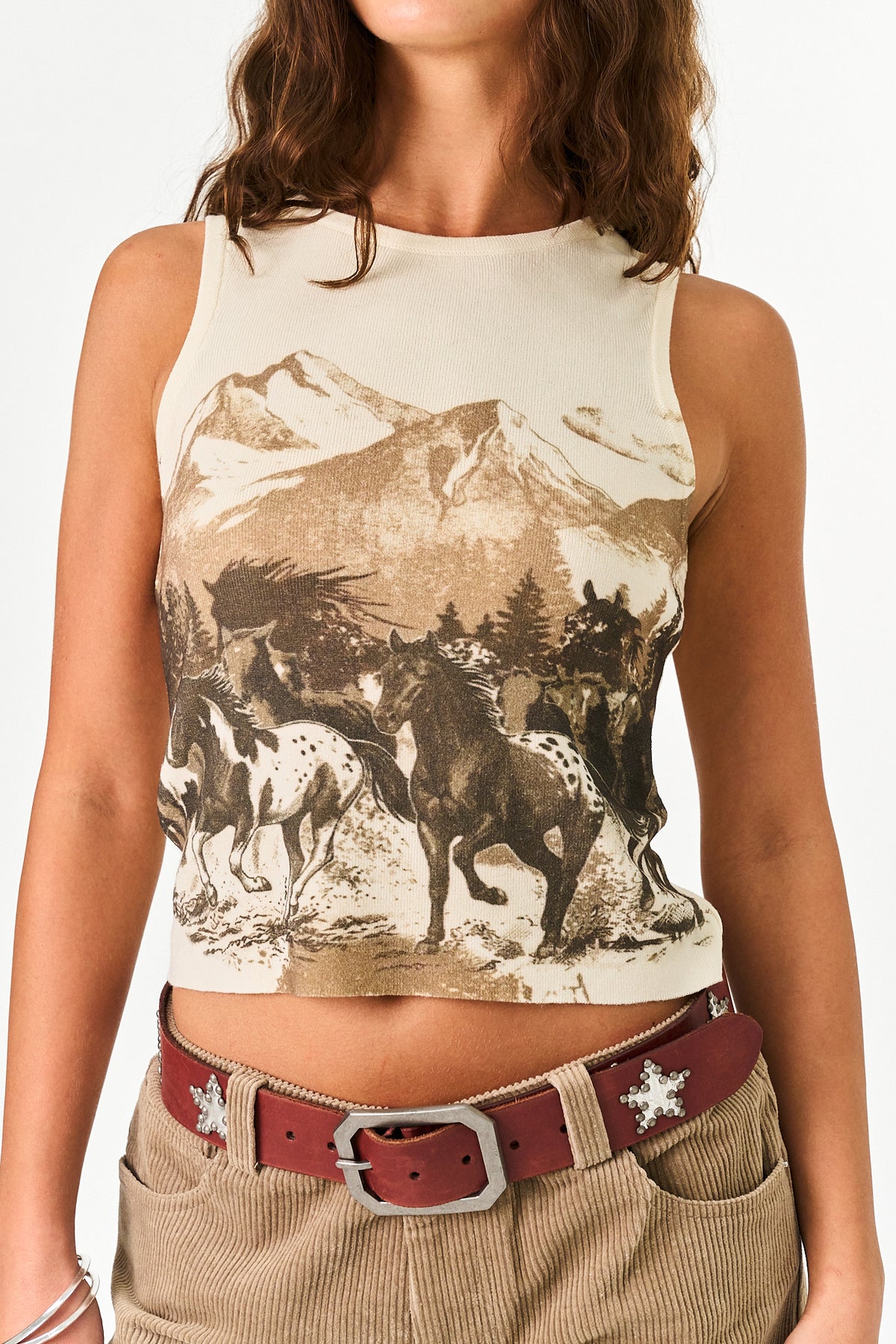 A woman wearing a sleeveless ribbed crop top with a brown and beige graphic print of wild horses in front of mountains, paired with khaki pants and a red belt.