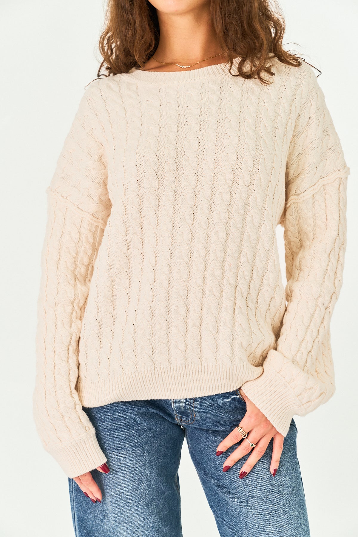 A woman wearing a cream-colored cable-knit sweater and blue jeans, shown from the neck down against a plain white background.