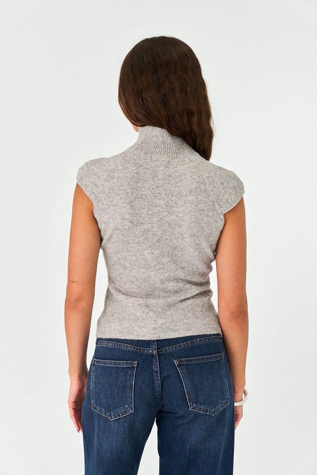 A woman viewed from behind wearing a light gray heathered knit mock-neck top with cap sleeves and dark blue jeans, set against a plain white studio background.