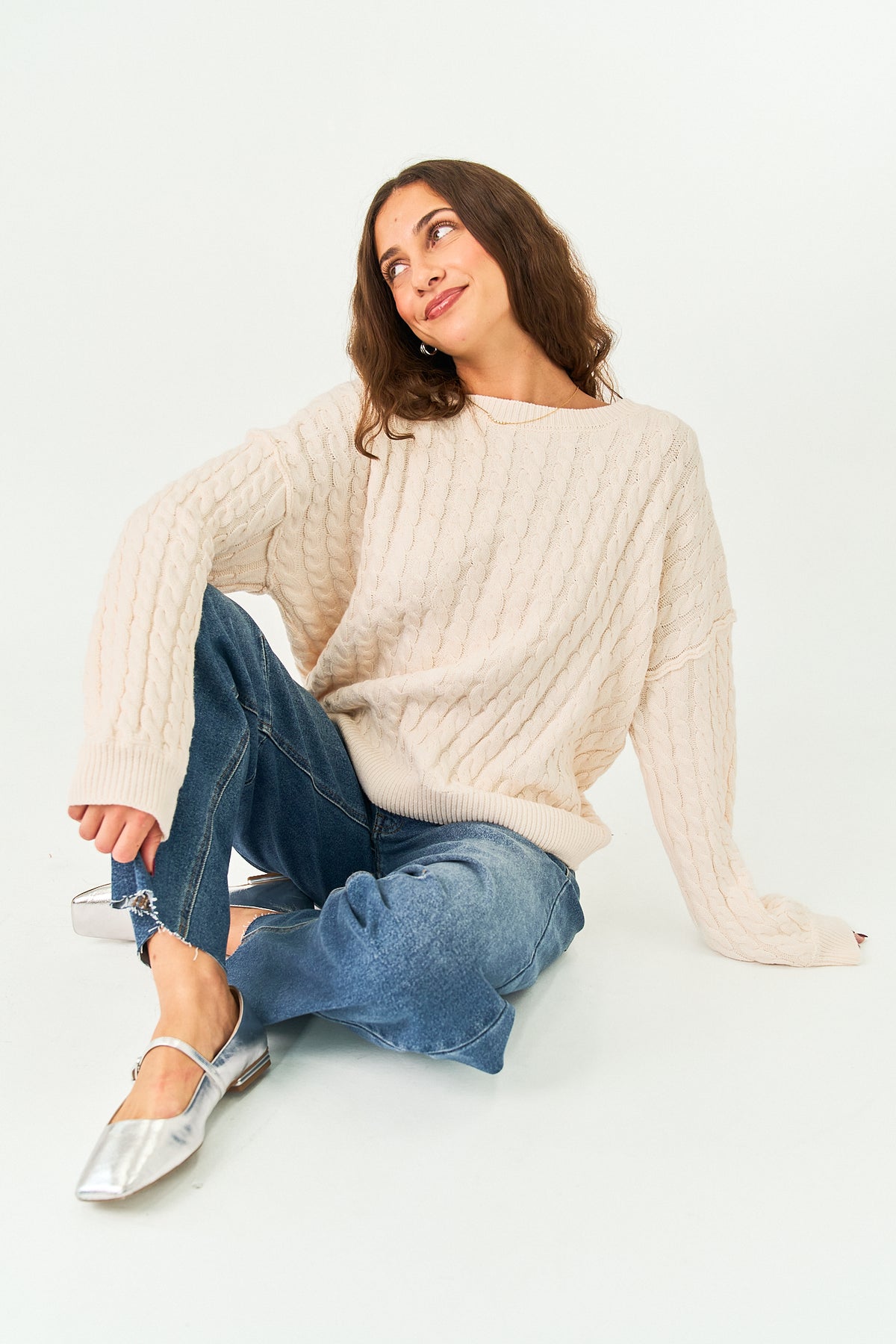 A woman sits on a white studio floor wearing a cream cable-knit sweater, blue jeans, and silver flat shoes.