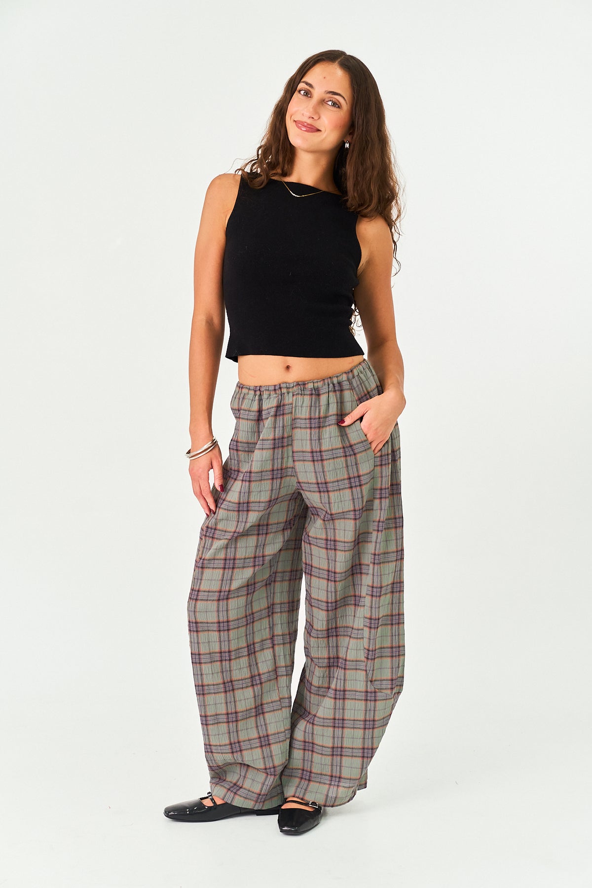 A woman modeling a pair of wide-leg plaid trousers in a gray, green, and red tartan pattern, paired with a black sleeveless crop top and black flats, against a plain white studio background.