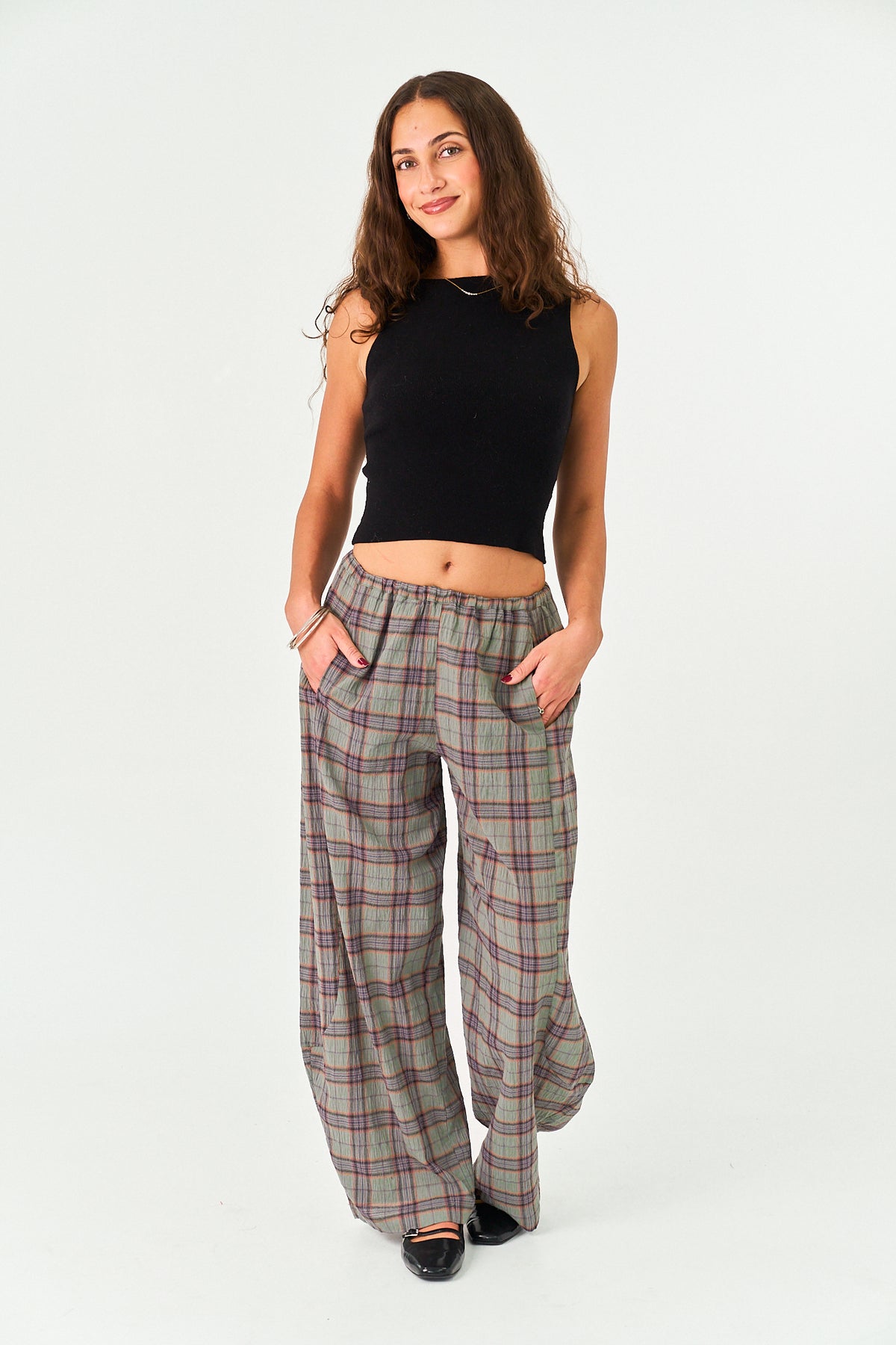 A woman modeling a pair of wide-leg plaid trousers in a gray, green, and red tartan pattern, paired with a black sleeveless crop top, against a plain white studio background.