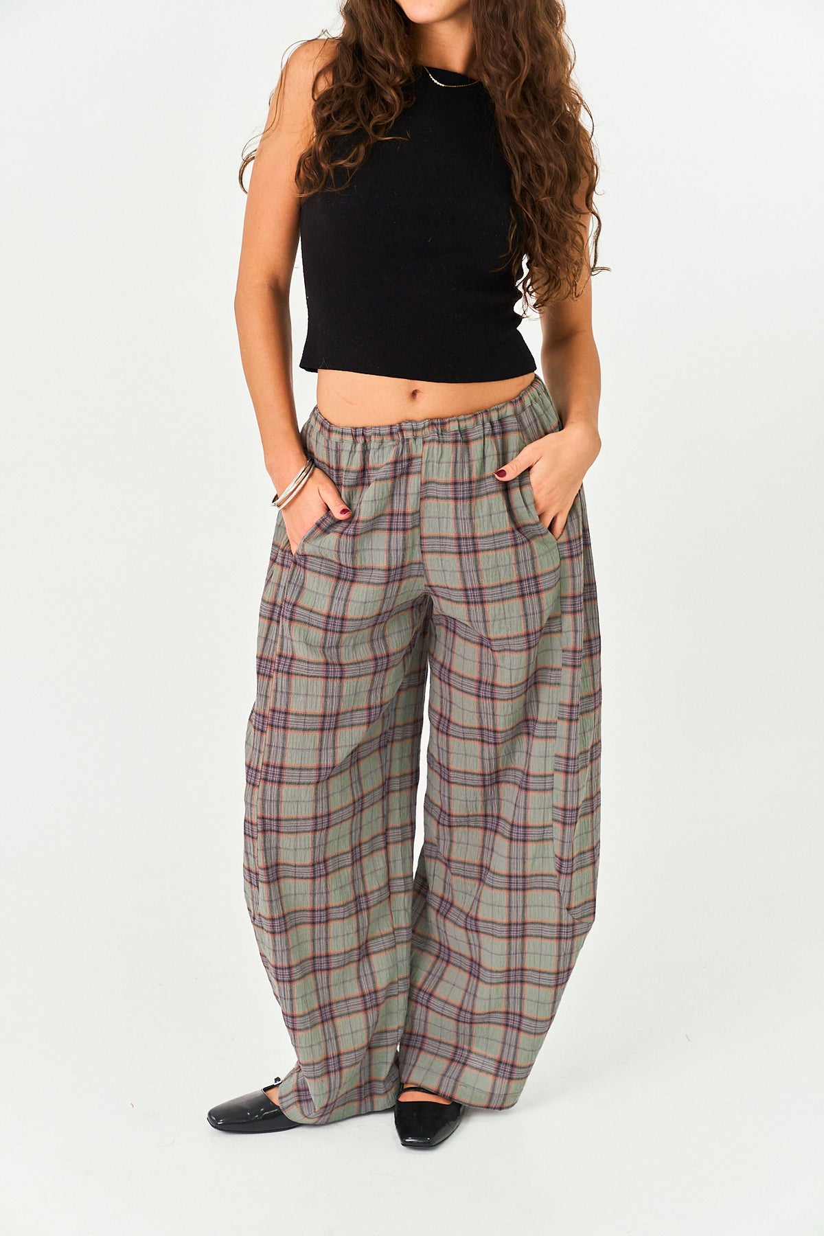 A model wearing a pair of wide-leg plaid trousers in a muted green-gray with red and blue lines, paired with a black sleeveless crop top and black flats, against a plain white studio background.