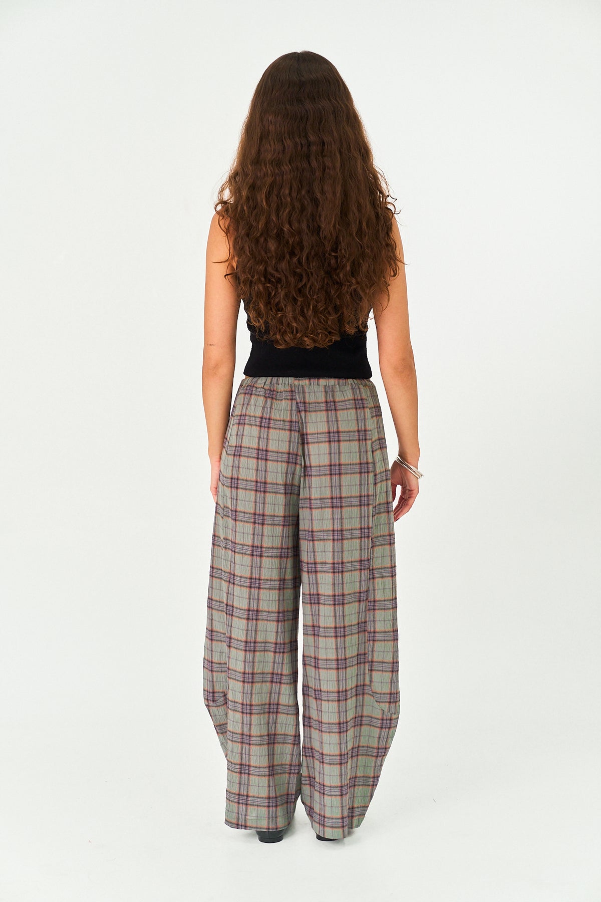A woman with long, curly brown hair is shown from behind wearing wide-leg plaid trousers in a beige, gray, and red pattern, paired with a black sleeveless top, against a plain white studio background.