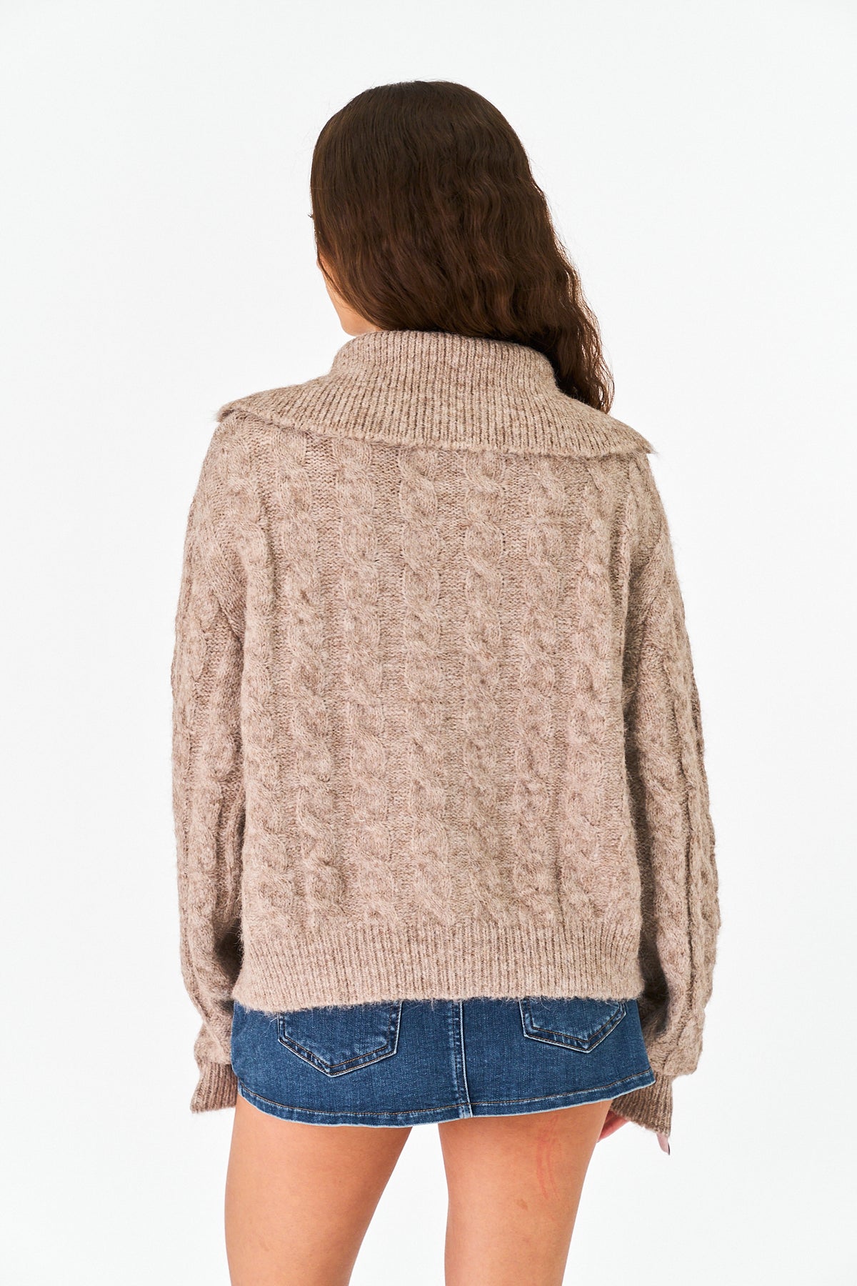 A woman viewed from behind wearing a chunky, textured knit sweater in a warm beige color, paired with a blue denim mini skirt, against a plain white background.