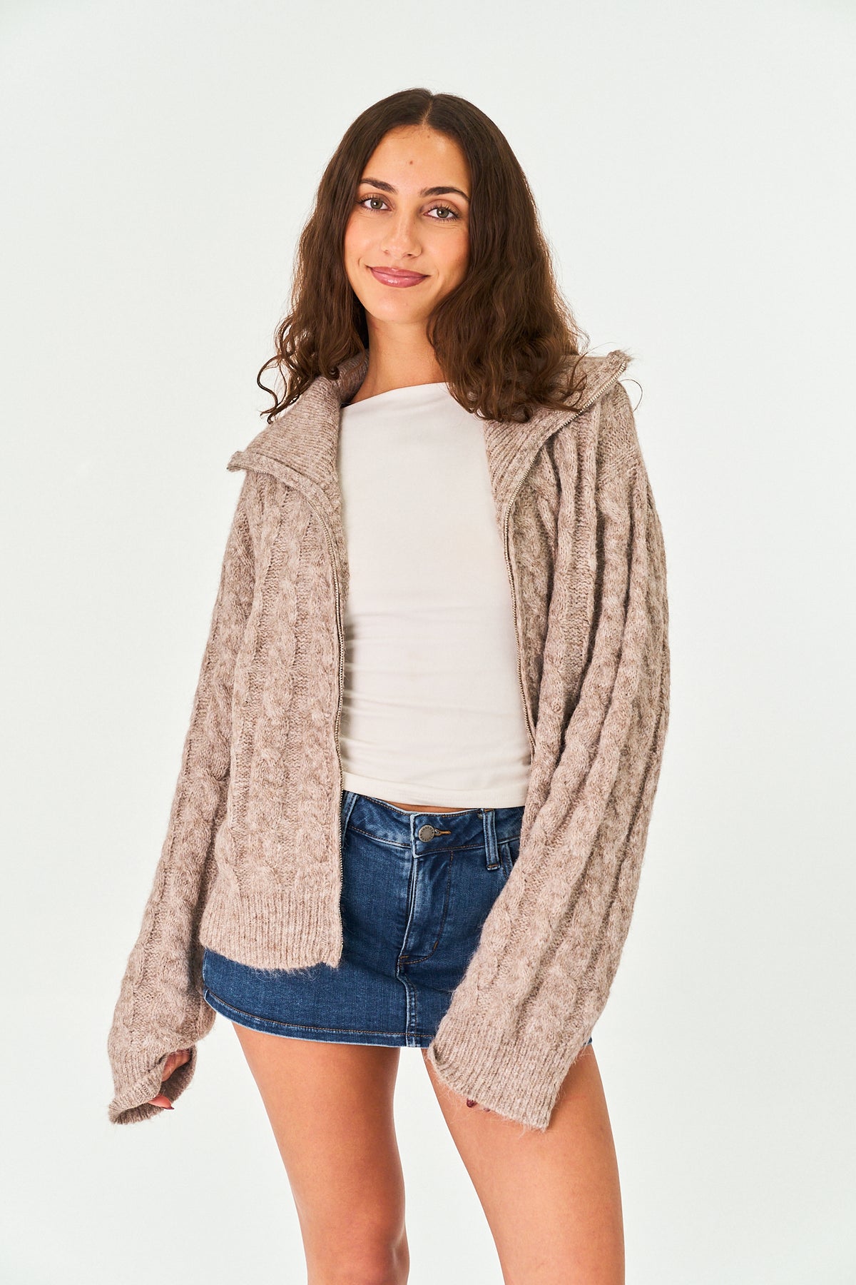 A woman modeling a chunky, oversized knit cardigan in a heathered taupe-beige color, worn open over a white top and blue denim shorts, against a plain white studio background.