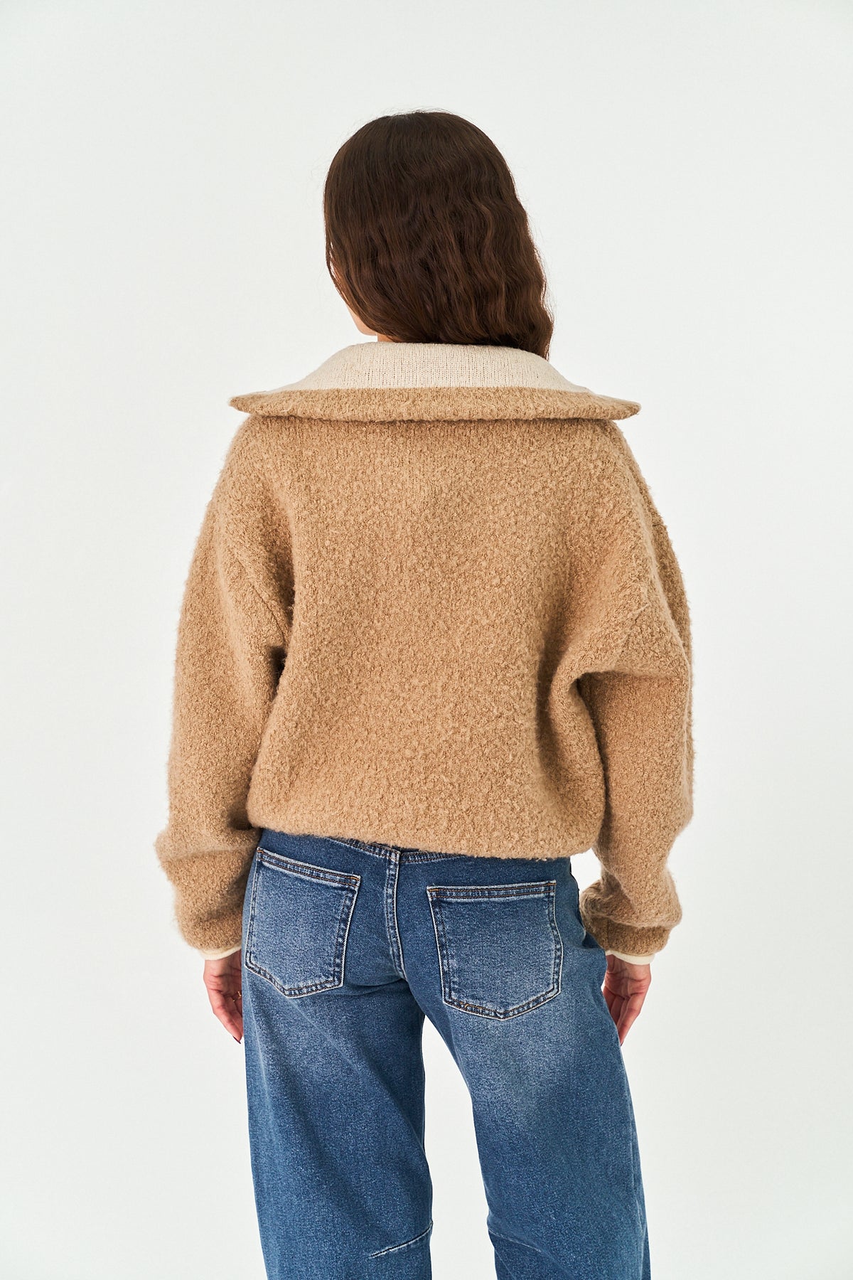 A person shown from the back wearing a cropped, fuzzy tan teddy-bear style jacket with a wide collar and blue jeans, against a plain light background.