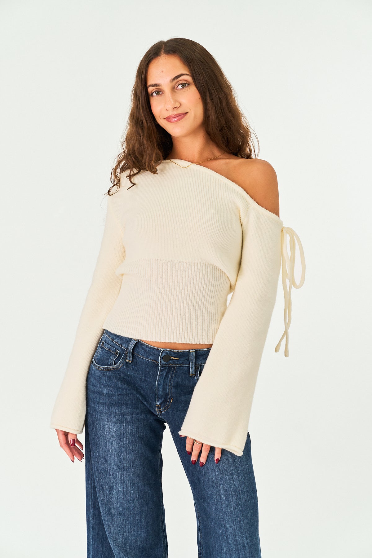 A woman modeling a cream off-the-shoulder knit sweater with long bell sleeves and side tie details, paired with blue jeans, against a plain white studio background.