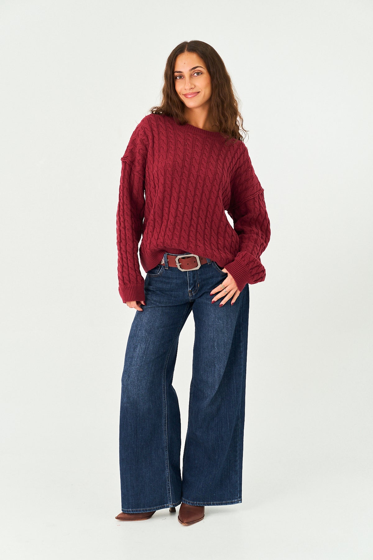 A woman modeling a chunky cable-knit sweater in deep burgundy, paired with wide-leg blue jeans, a brown belt, and brown boots, against a plain white studio background.
