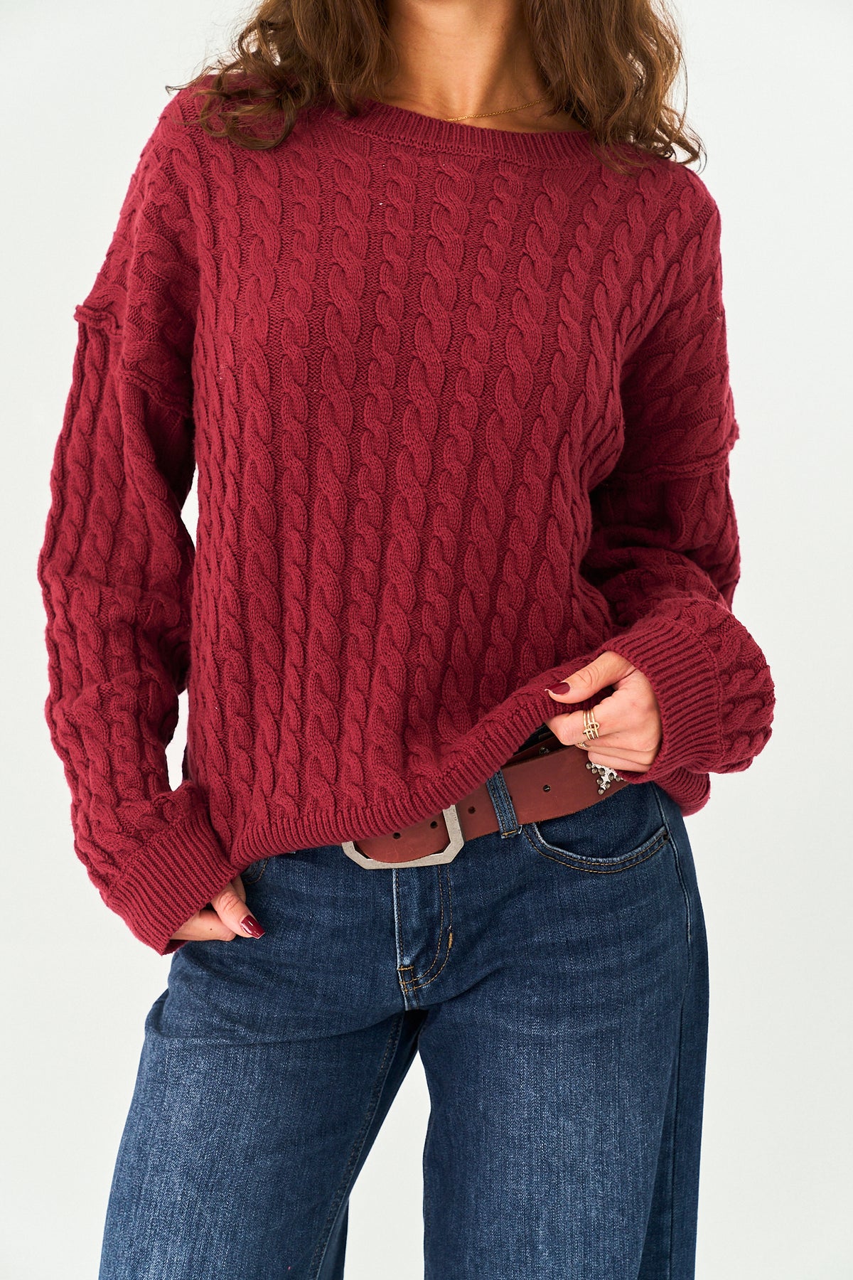 A woman wearing a chunky cable-knit sweater in a deep burgundy red, paired with blue jeans and a brown belt, against a plain white background.