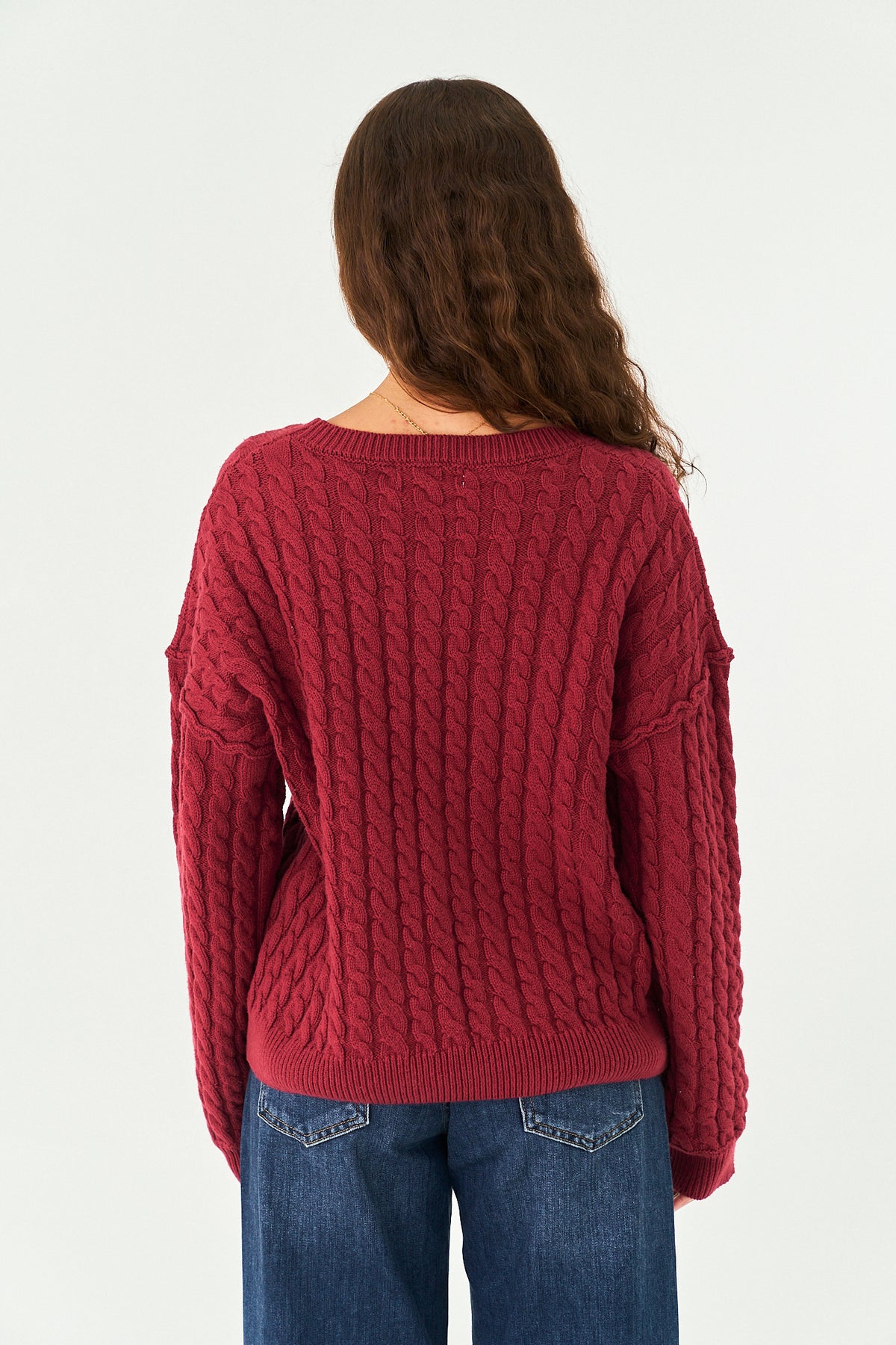 A woman viewed from the back wearing a chunky cable-knit sweater in a deep burgundy red, paired with blue jeans, against a plain white studio background.