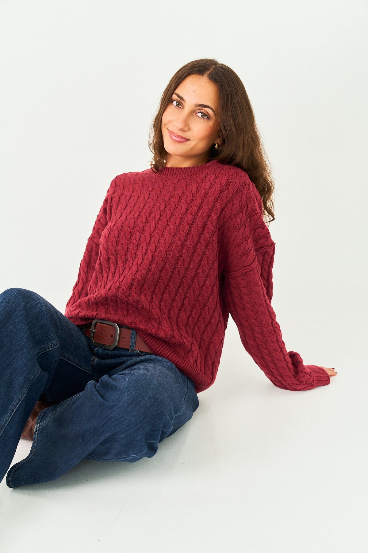A woman wearing a chunky cable-knit sweater in burgundy and blue jeans, sitting against a plain white studio background.