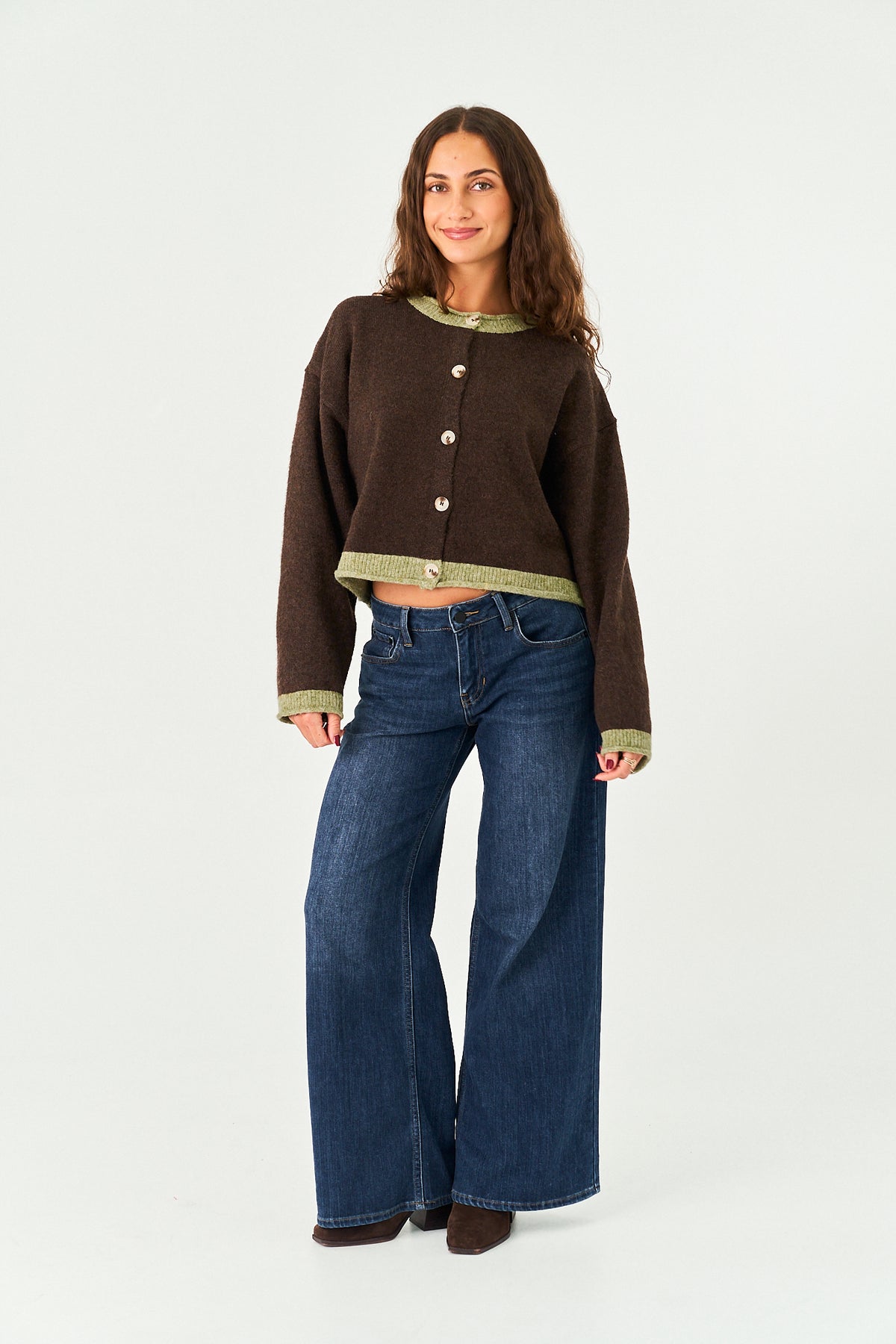 A woman modeling a dark brown cropped cardigan with olive green trim and buttons, paired with wide-leg blue jeans, against a plain white studio background.